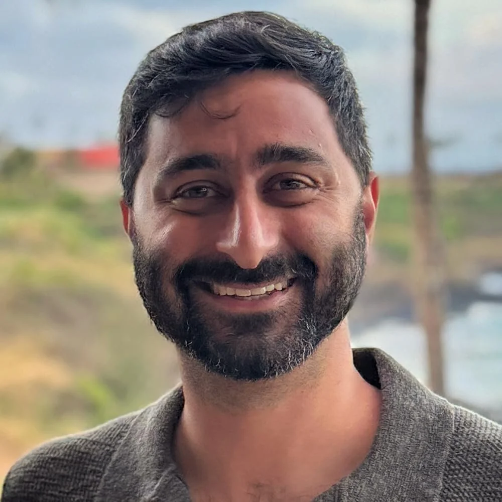 A smiling man with dark hair and a beard, wearing a gray shirt, outdoors with a blurred background of trees and water.