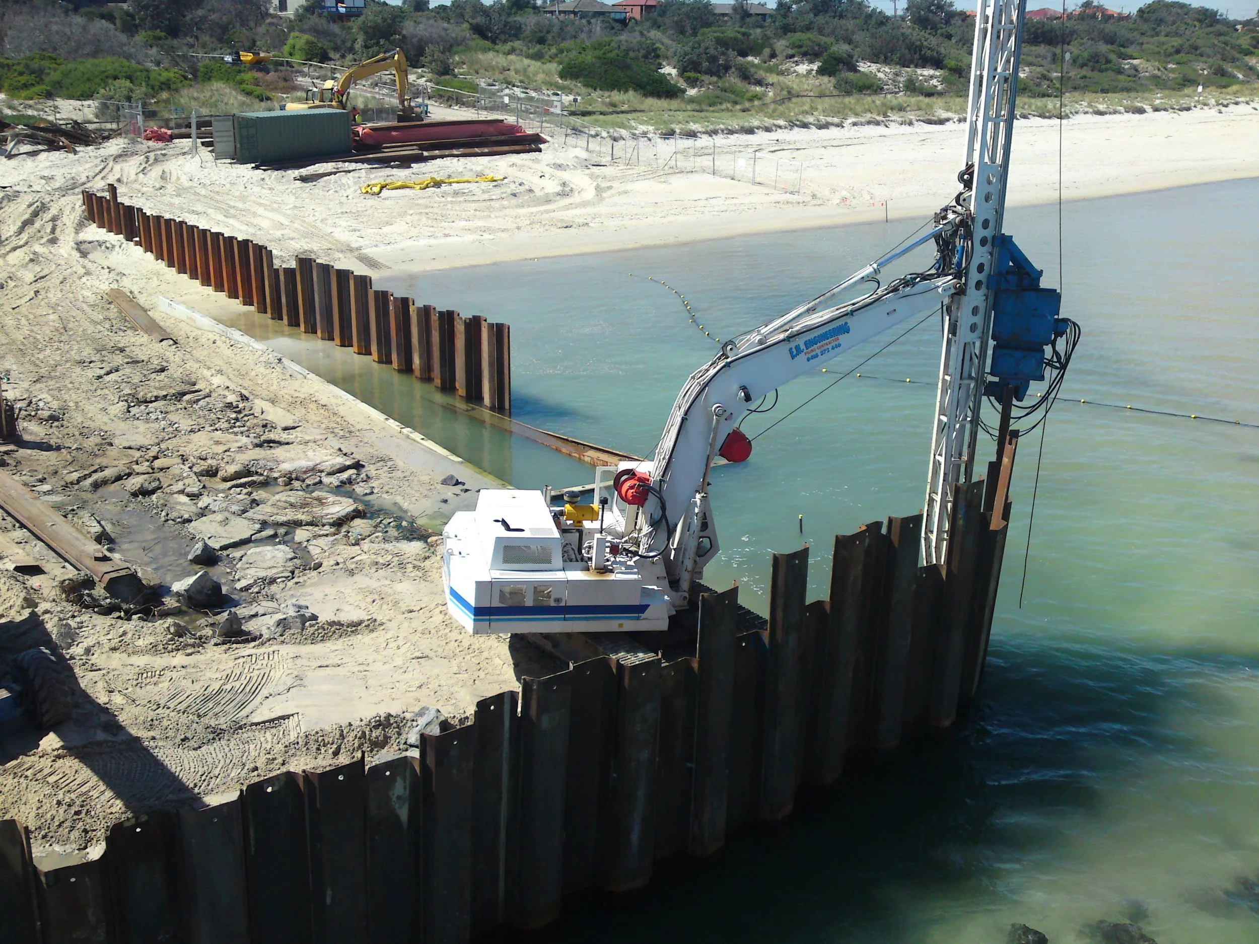 Construction equipment along the shoreline, with a large crane extending over the water, topped by a series of rusted steel sheet piles being driven into the ground for shoreline reinforcement.