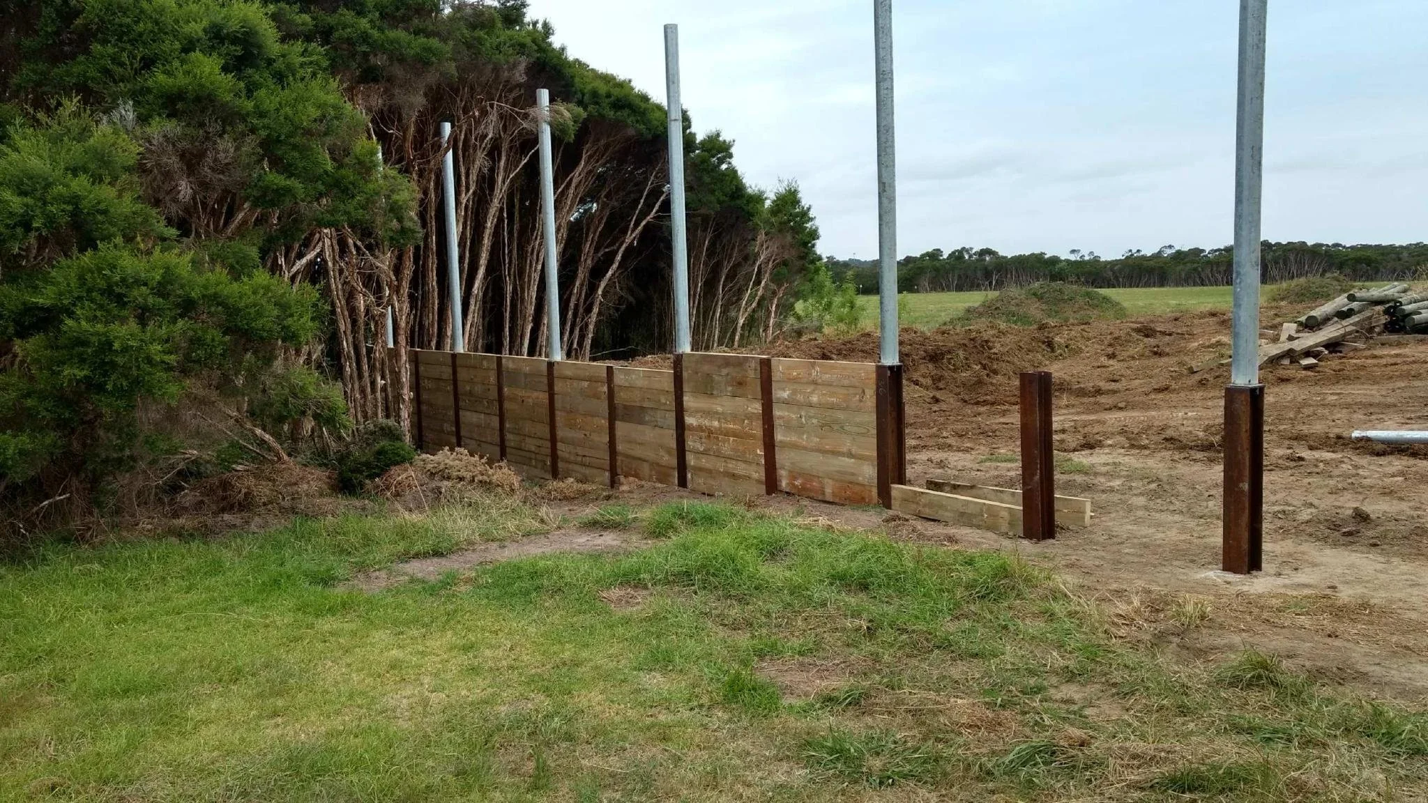Construction of a wooden and metal fence in a rural area, with trees and open fields in the background.