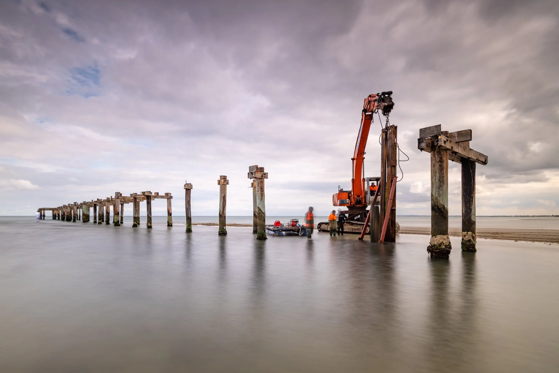 Construction workers using a crane to restore a wooden pier over the ocean on a cloudy day.