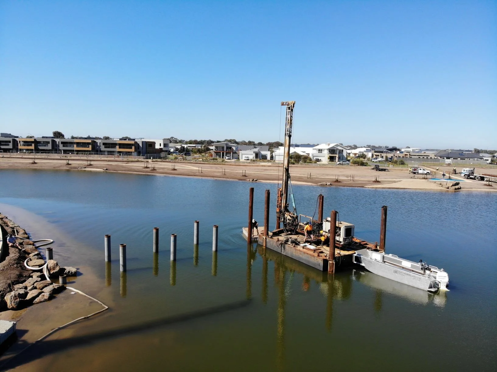 Construction barge with a crane on water, surrounded by pilings, near a sandy shore and residential buildings in the background under a clear blue sky.