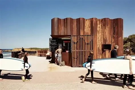 People carrying surfboards outside a small building with a rusted metal exterior on a beach walkway, under a clear blue sky.