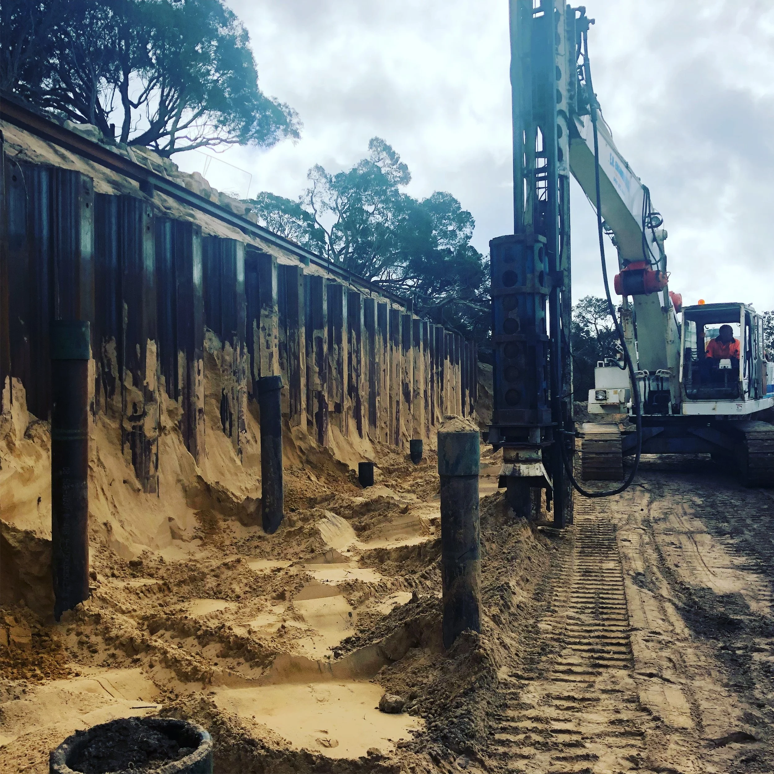 Construction site with a large excavator drilling into the ground near a sloped dirt wall reinforced with metal sheets, with trees and cloudy sky in the background.