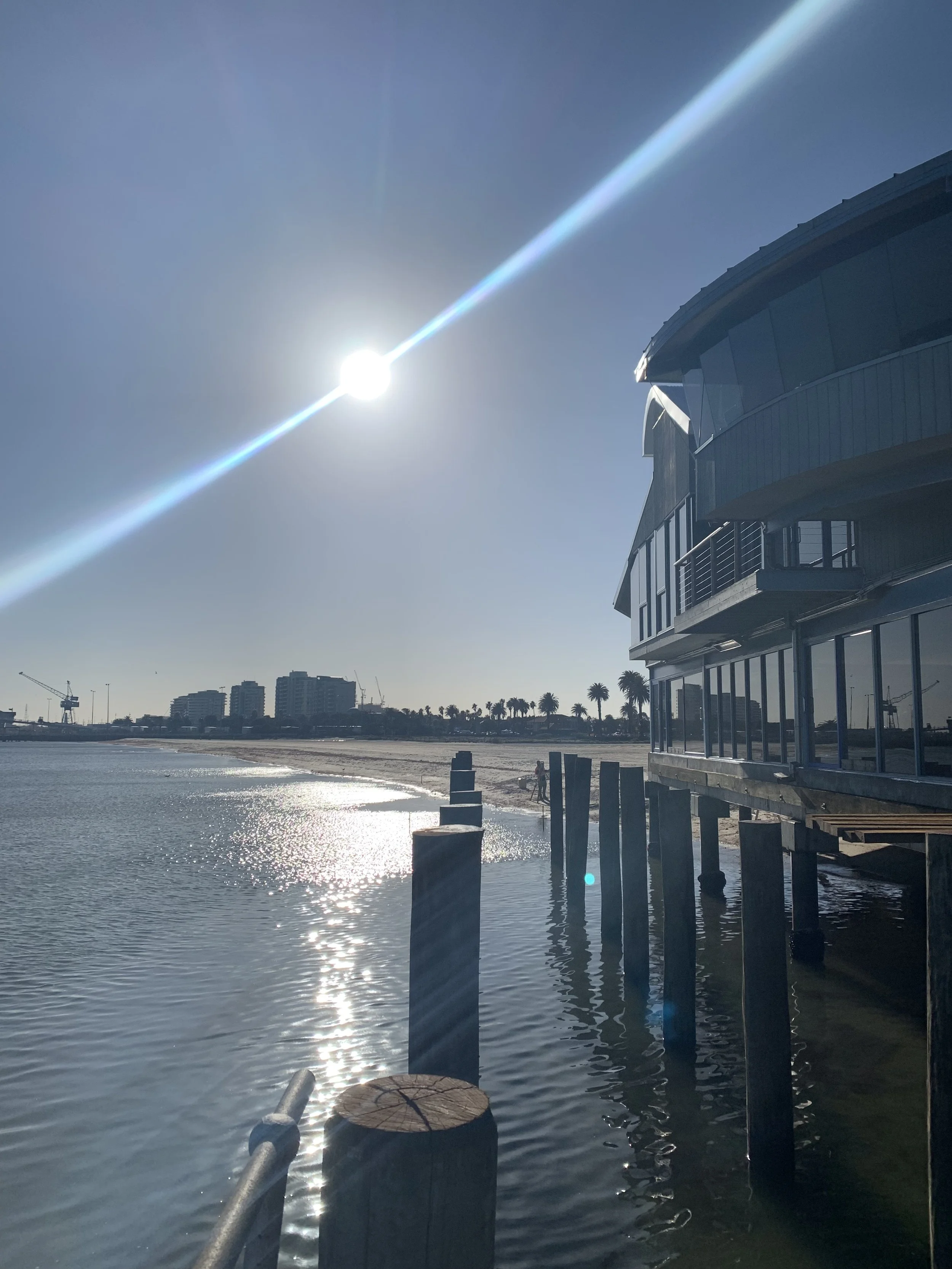 A waterfront scene with the sun shining brightly in the sky, reflecting off the water, alongside a modern building on the right, a sandy shoreline, and city buildings in the background.