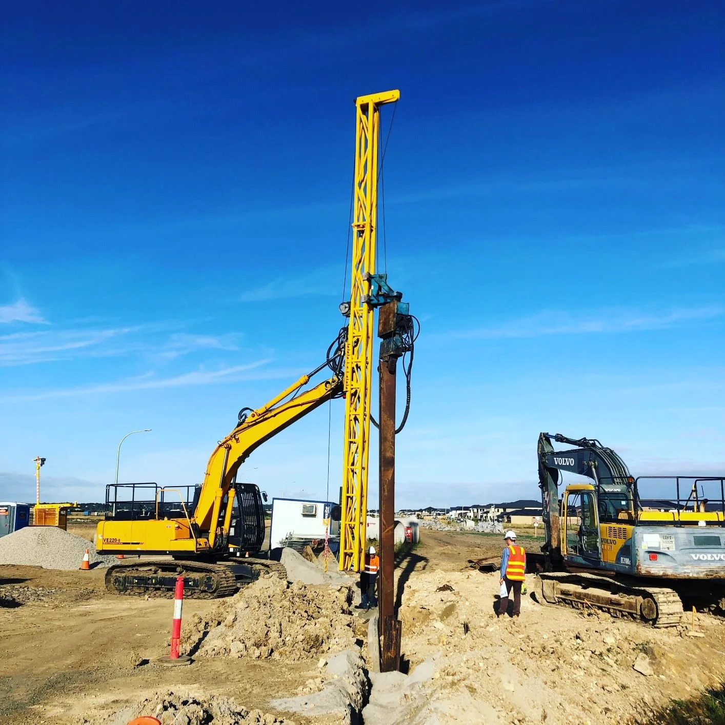 Construction site with two excavators, one yellow and one black, digging in the dirt. A worker wearing a hard hat and safety vest stands near the excavators. A large blue sky and distant houses are visible in the background.