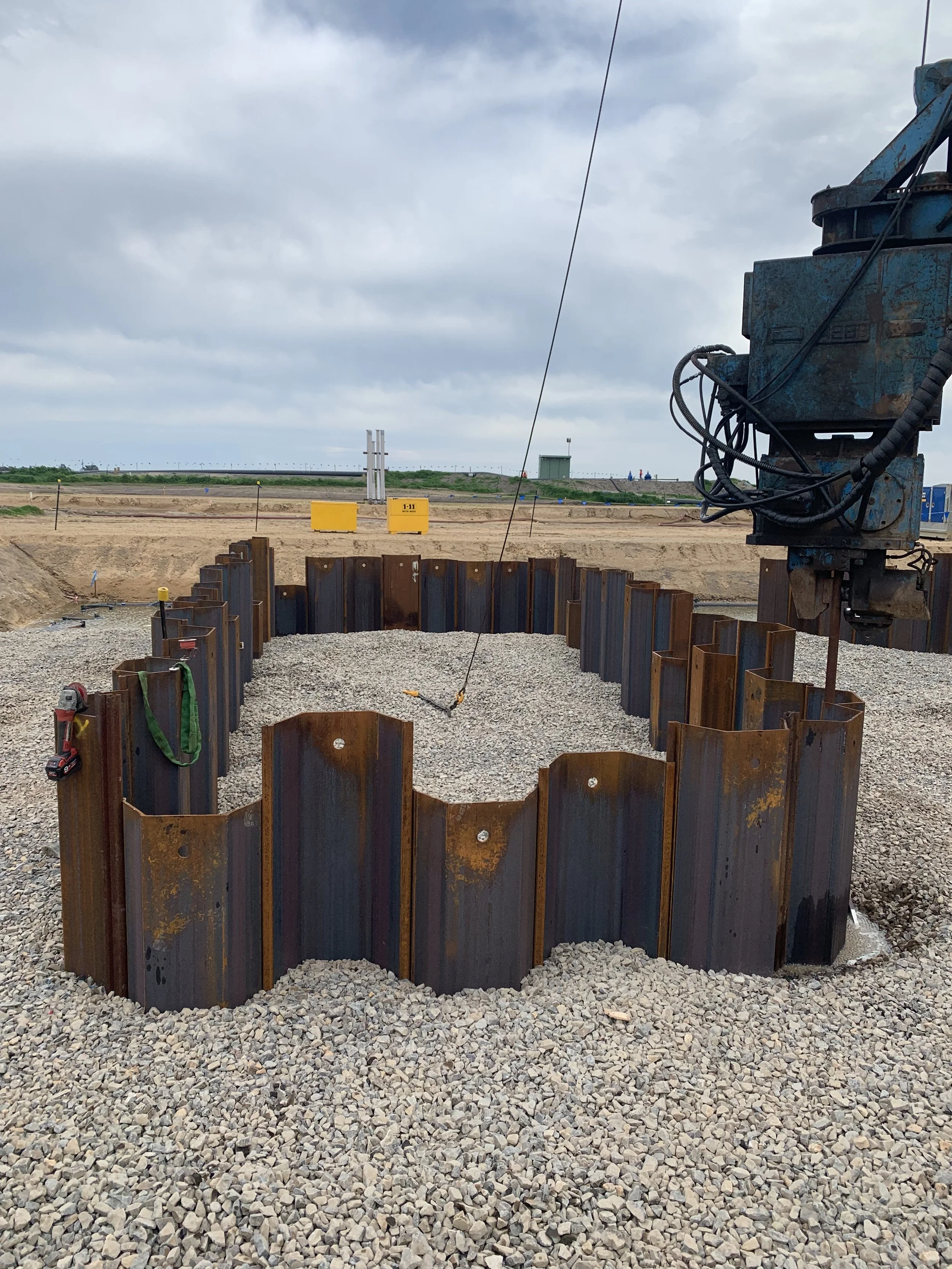 Construction site with steel sheet pile walls forming a circular excavation, gravel covering the bottom, and construction equipment present; cloudy sky in the background.
