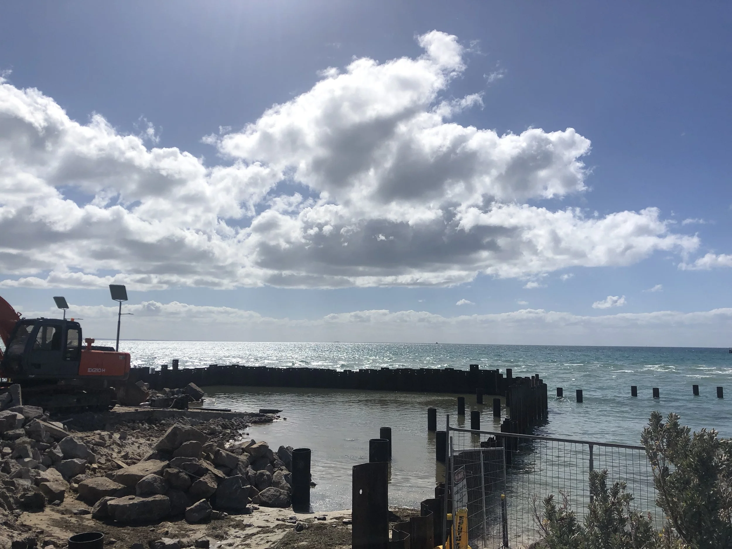 A coastal scene with construction equipment on a rocky shore, a partially built seawall, and a metal fence in the foreground. The ocean and a partly cloudy sky are in the background.
