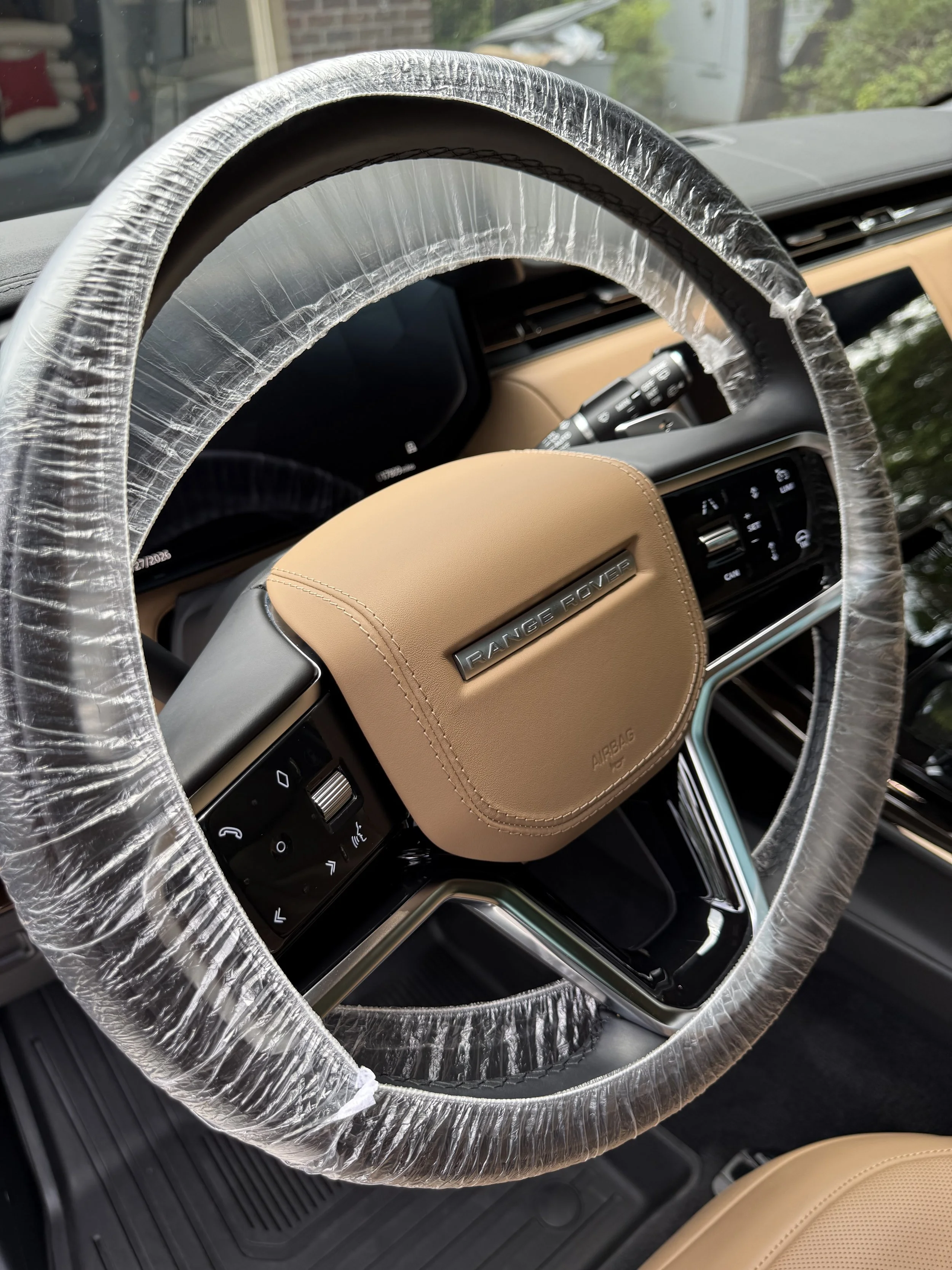Close-up of a Porsche Range Rover interior steering wheel wrapped in clear protective plastic covering, with dashboard and controls visible behind.