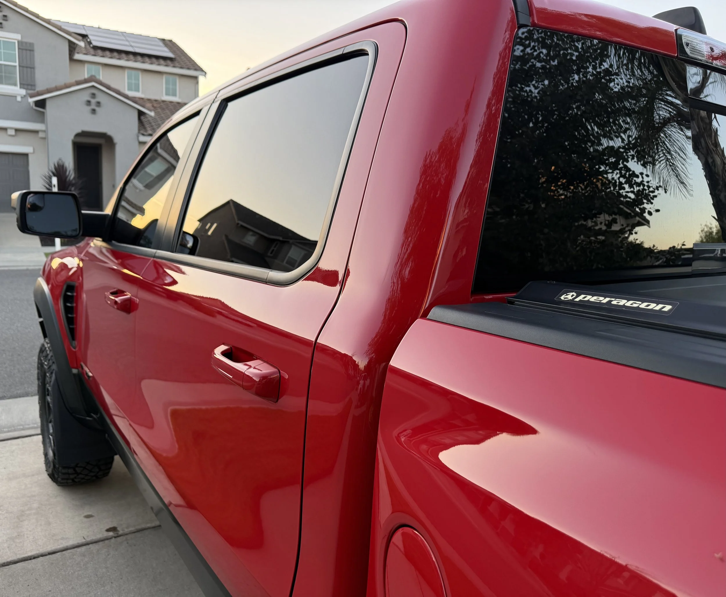 Close-up of a red pickup truck parked in a driveway in front of suburban houses, with a view of the side and rear windows reflecting nearby trees and sky.
