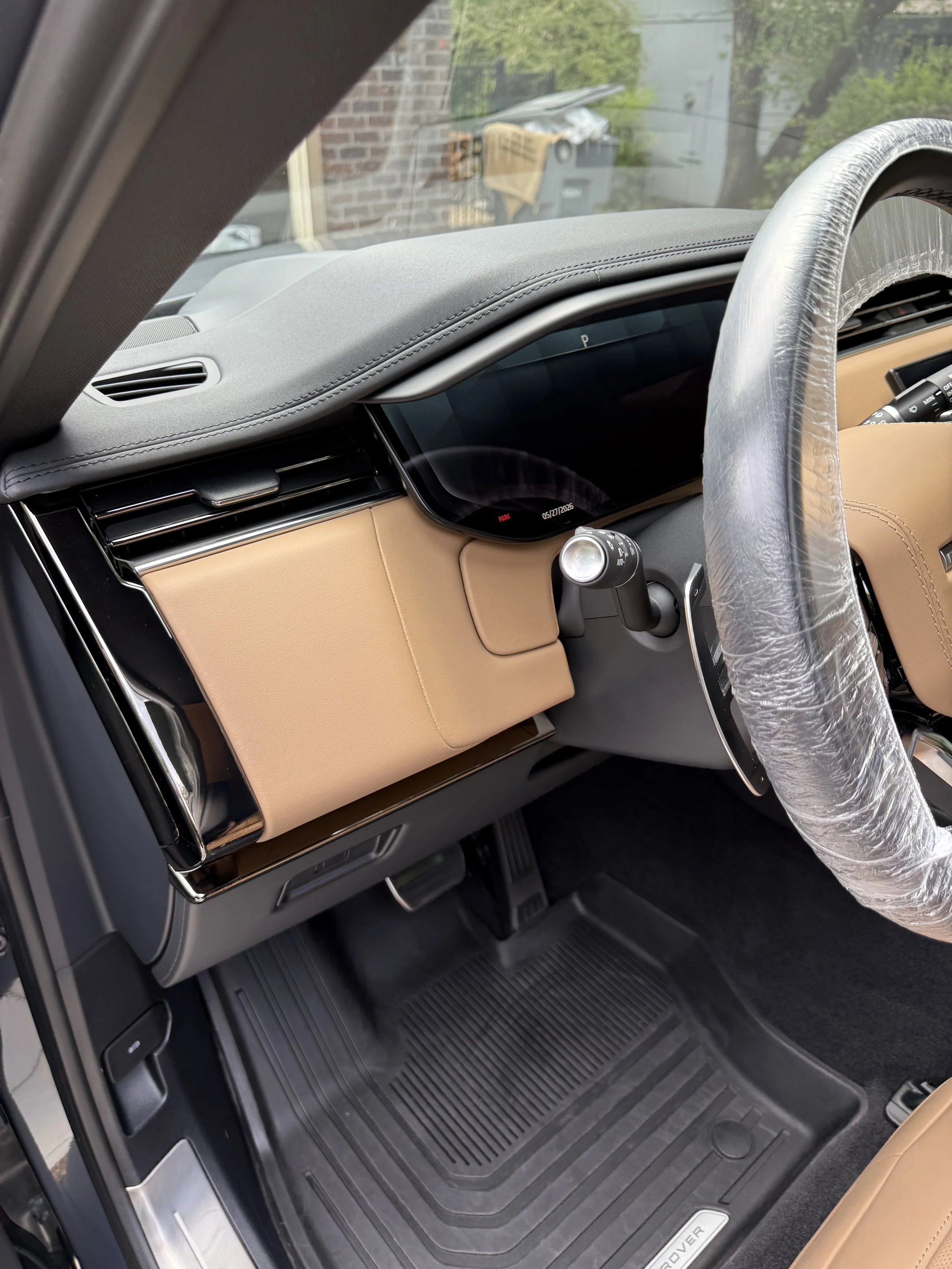 The interior of a luxury vehicle showing the dashboard, steering wheel covered in plastic, gear shift, and black floor mats with the car door open.