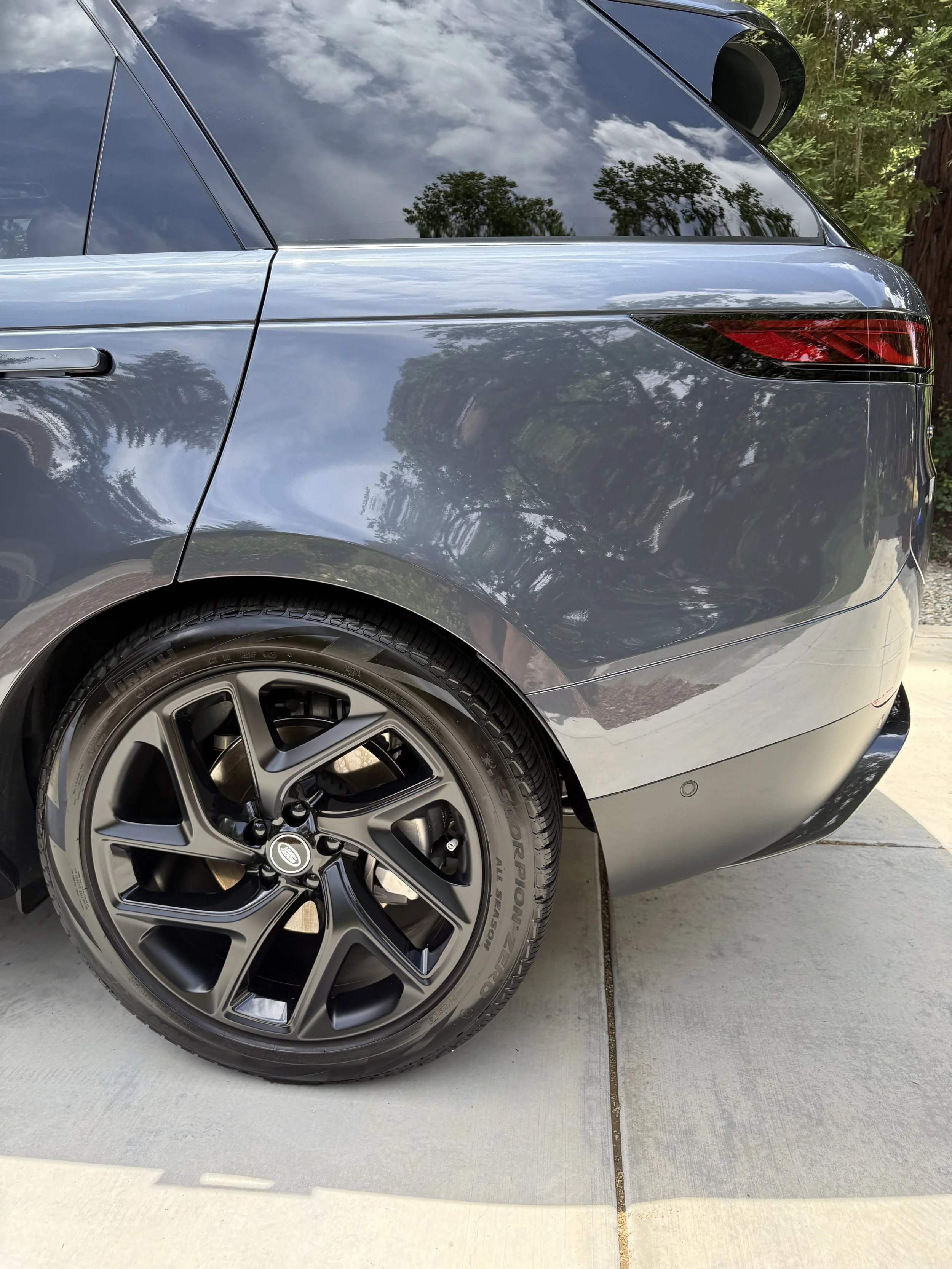 Close-up of the rear section of a dark gray modern car parked on concrete pavement next to some greenery, showing the wheel, tire, tail light, and part of the rear window with reflections of trees and clouds.