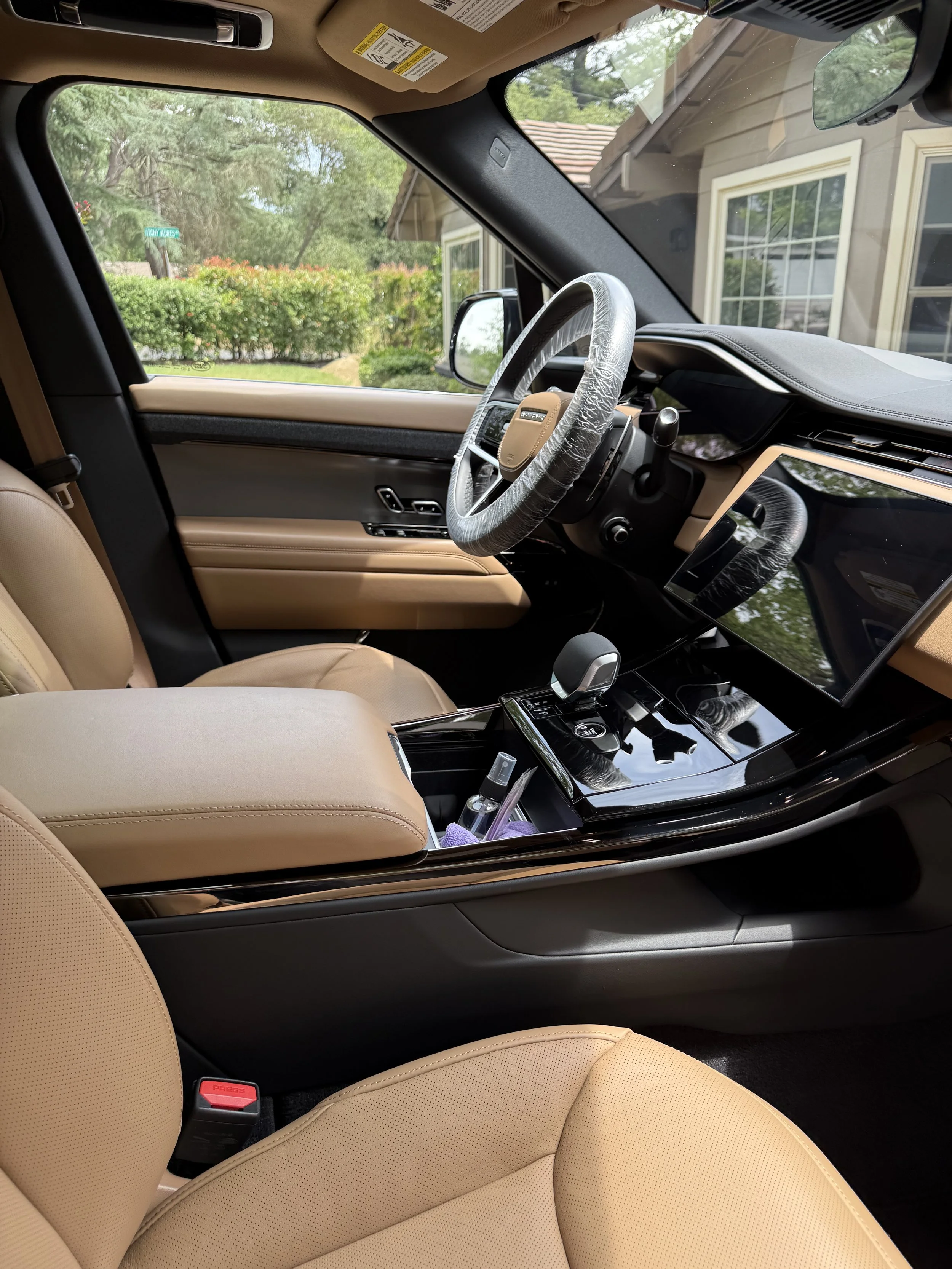 The interior of a car showing the front passenger seat, steering wheel with plastic cover, dashboard, and center console. The car's interior is beige with black accents. Outside the window, there is a house with windows and a garden with trees and bu