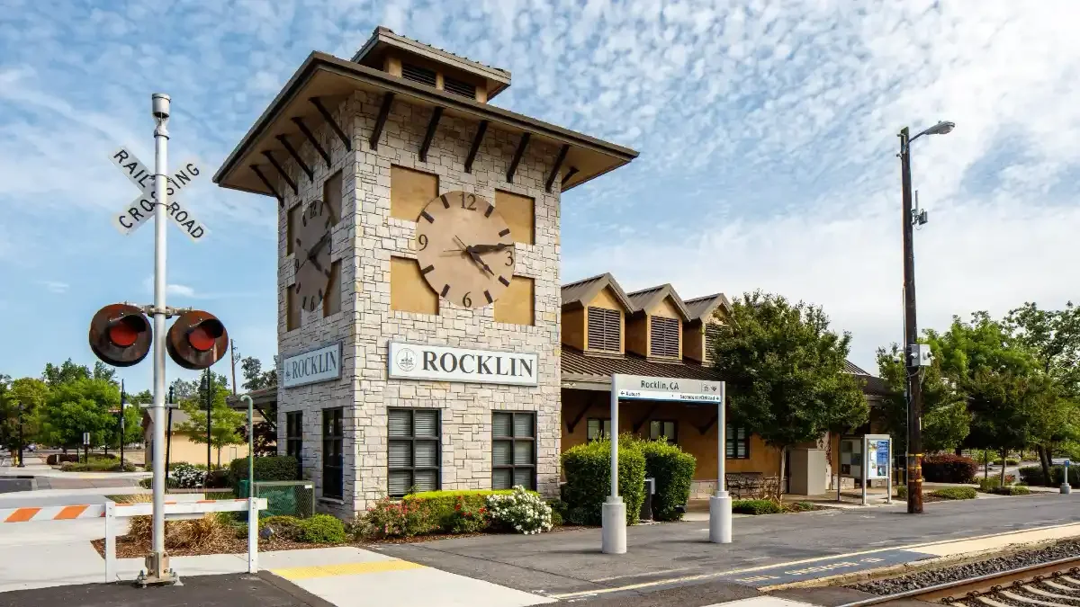 Clock tower at Rocklin train station in California with a railway crossing sign, benches, and trees in the background.