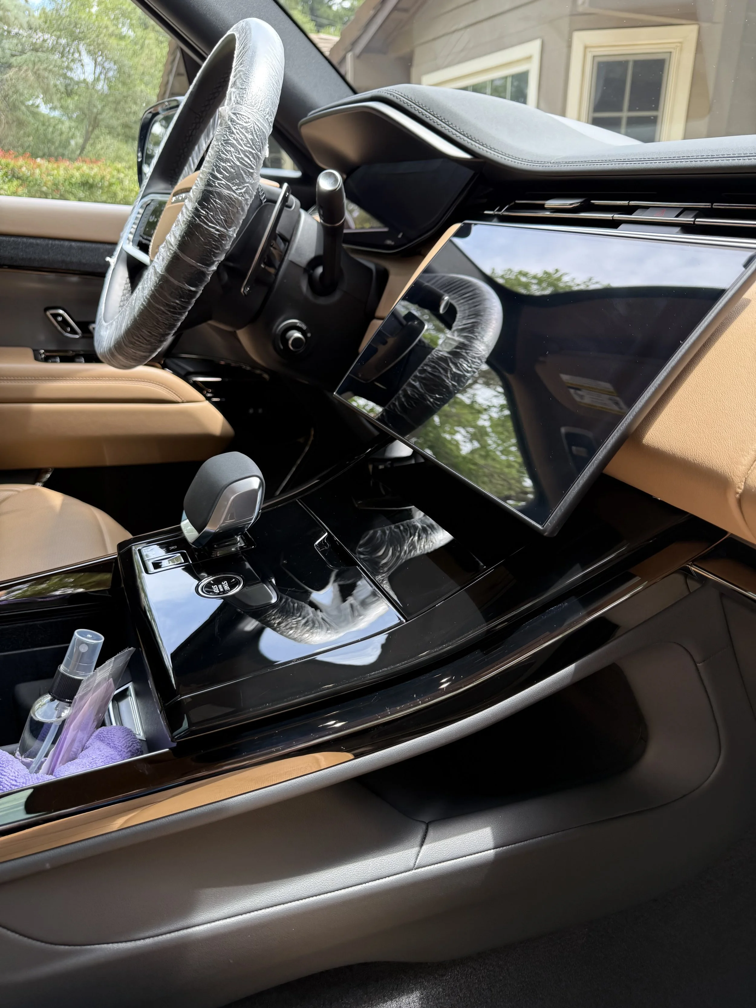The interior of a luxury car, showing the dashboard, steering wheel wrapped in plastic, center console with gear shift, and a large central touchscreen display with a black glossy finish. The door panel includes a control switch and a wood accent.