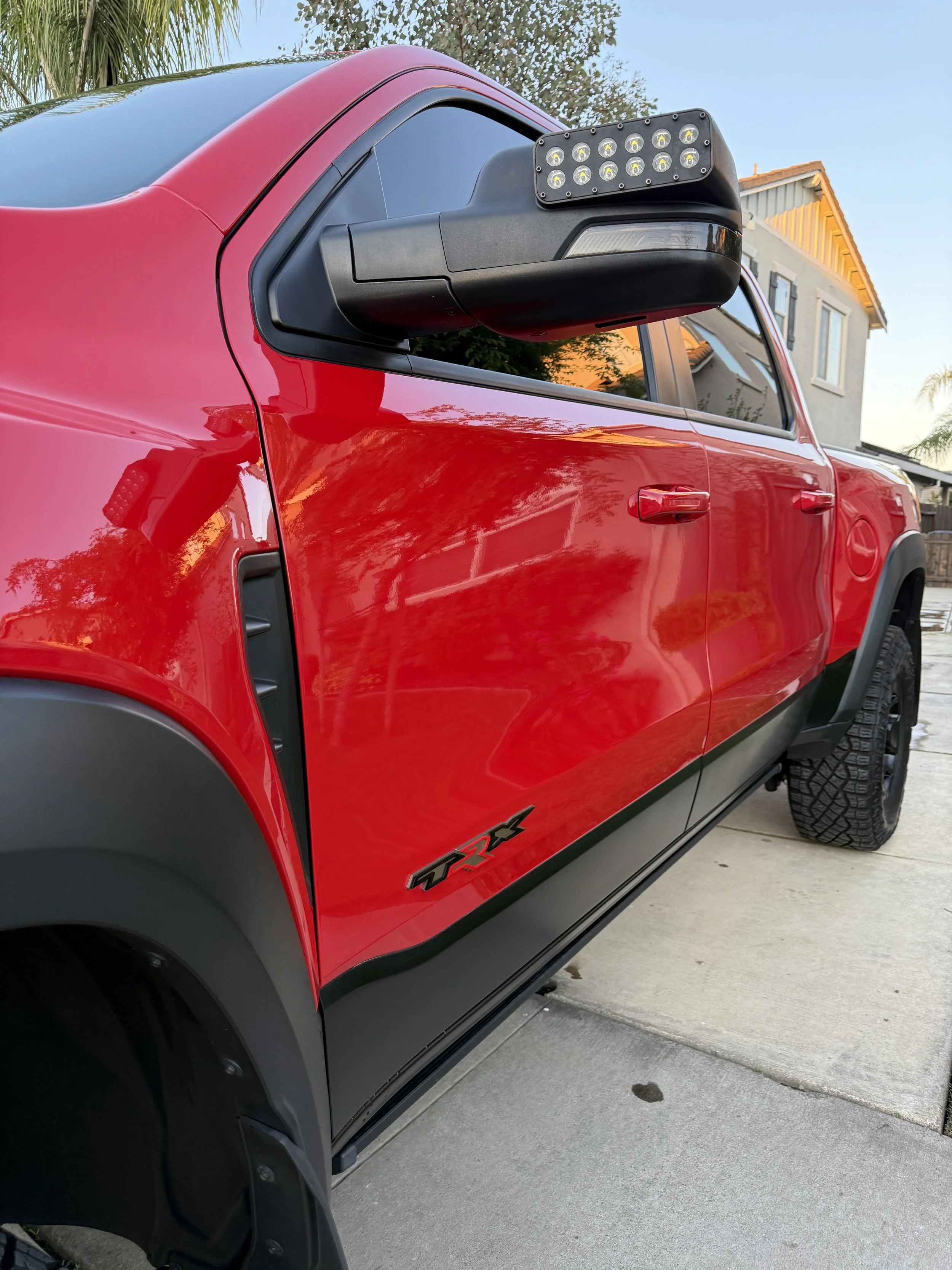 Rear side view of a red pickup truck with an illuminated light bar mounted on the side mirror, parked on a driveway near a residential house.