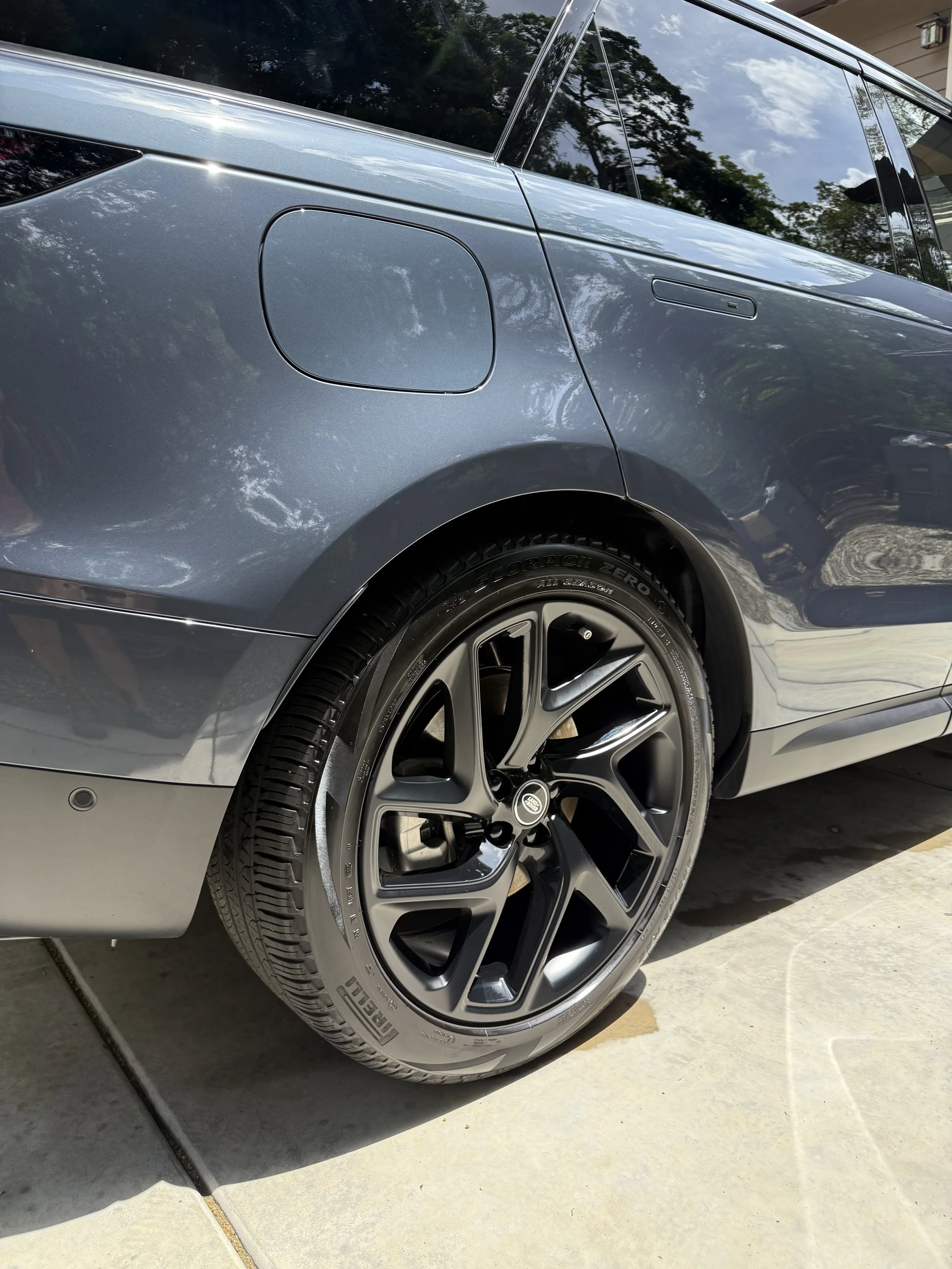 Close-up of a gray modern car showing part of the rear wheel, tire, fuel door, and the side windows, with reflections of trees and sky on the car's surface.