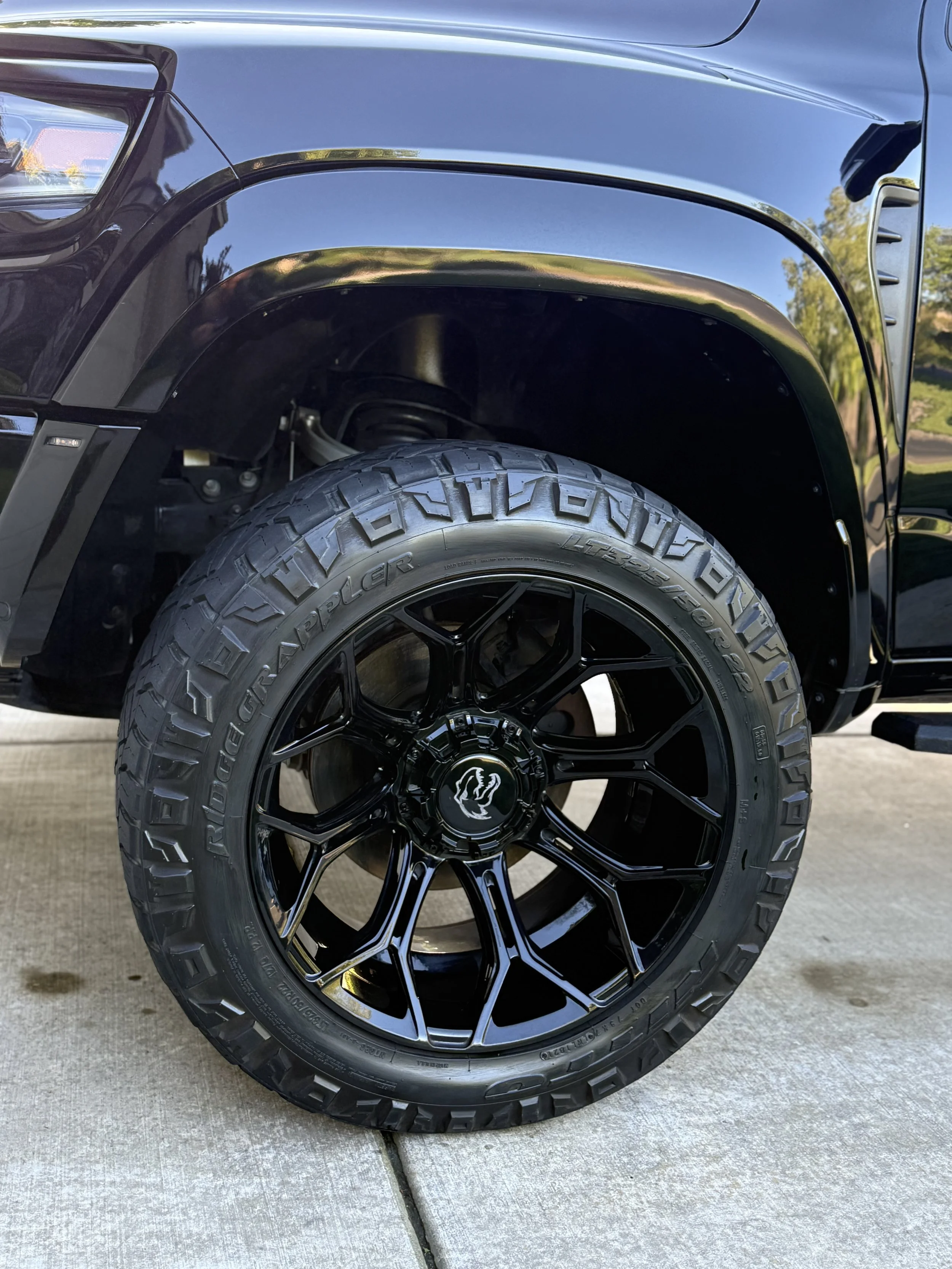 Close-up of a black vehicle's front tire with a large tread pattern and black alloy wheel, parked on a concrete driveway with greenery reflected on the glossy body.