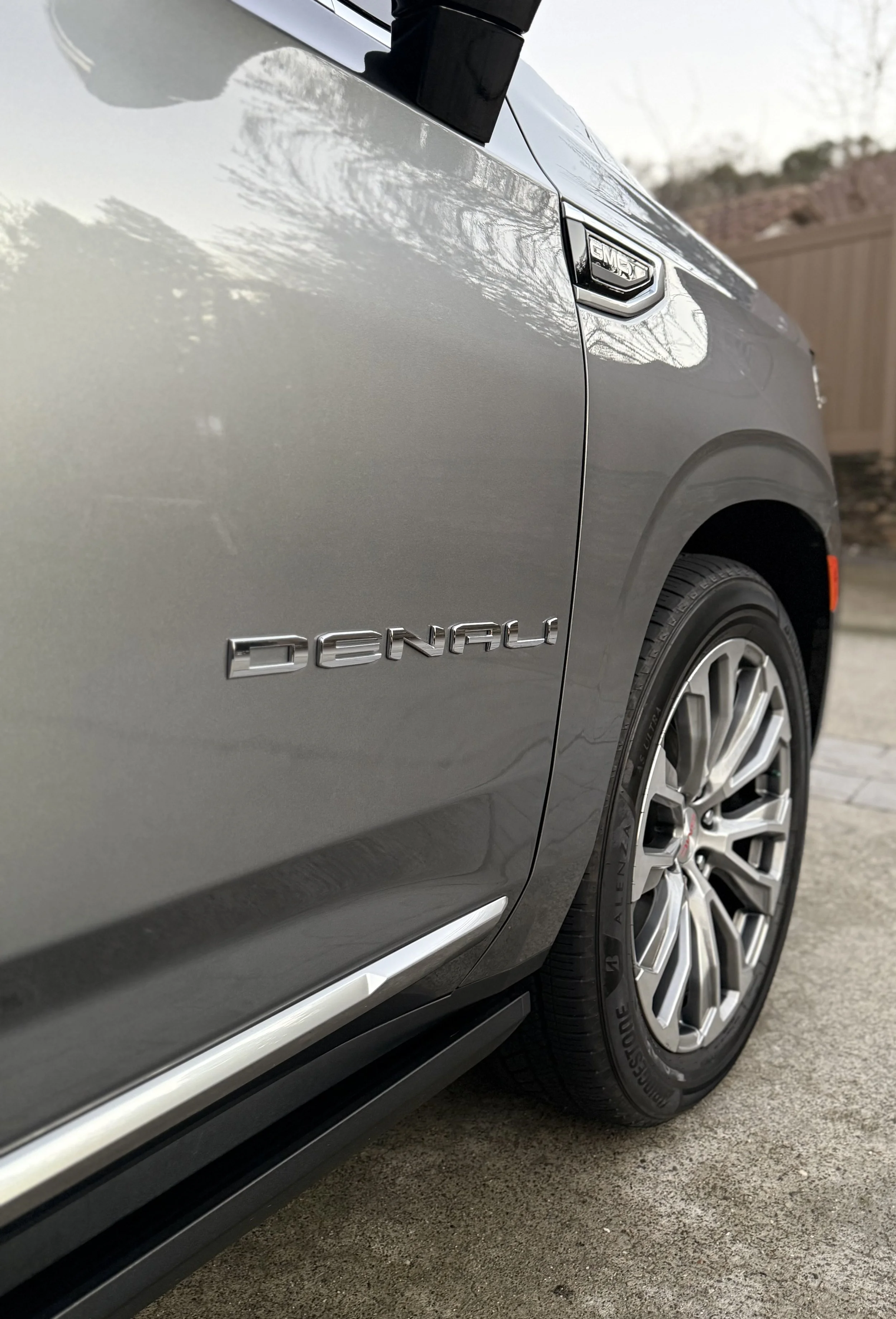 Close-up of the front side of a silver GMC Denali vehicle parked on a driveway, showing the Denali badge and part of the front wheel.