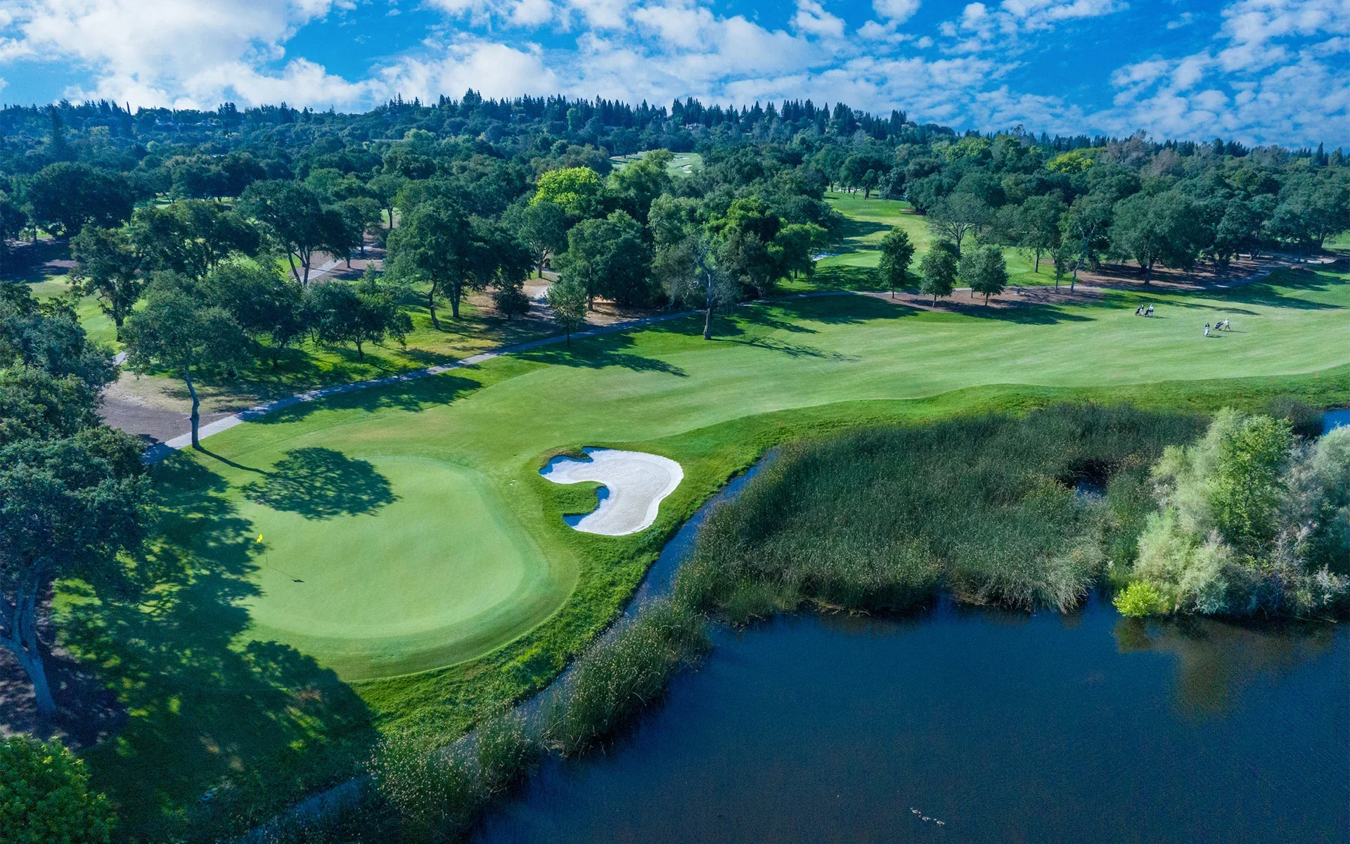Aerial view of a golf course with green fairways, water hazards, sand traps, and tall trees under a partly cloudy sky.