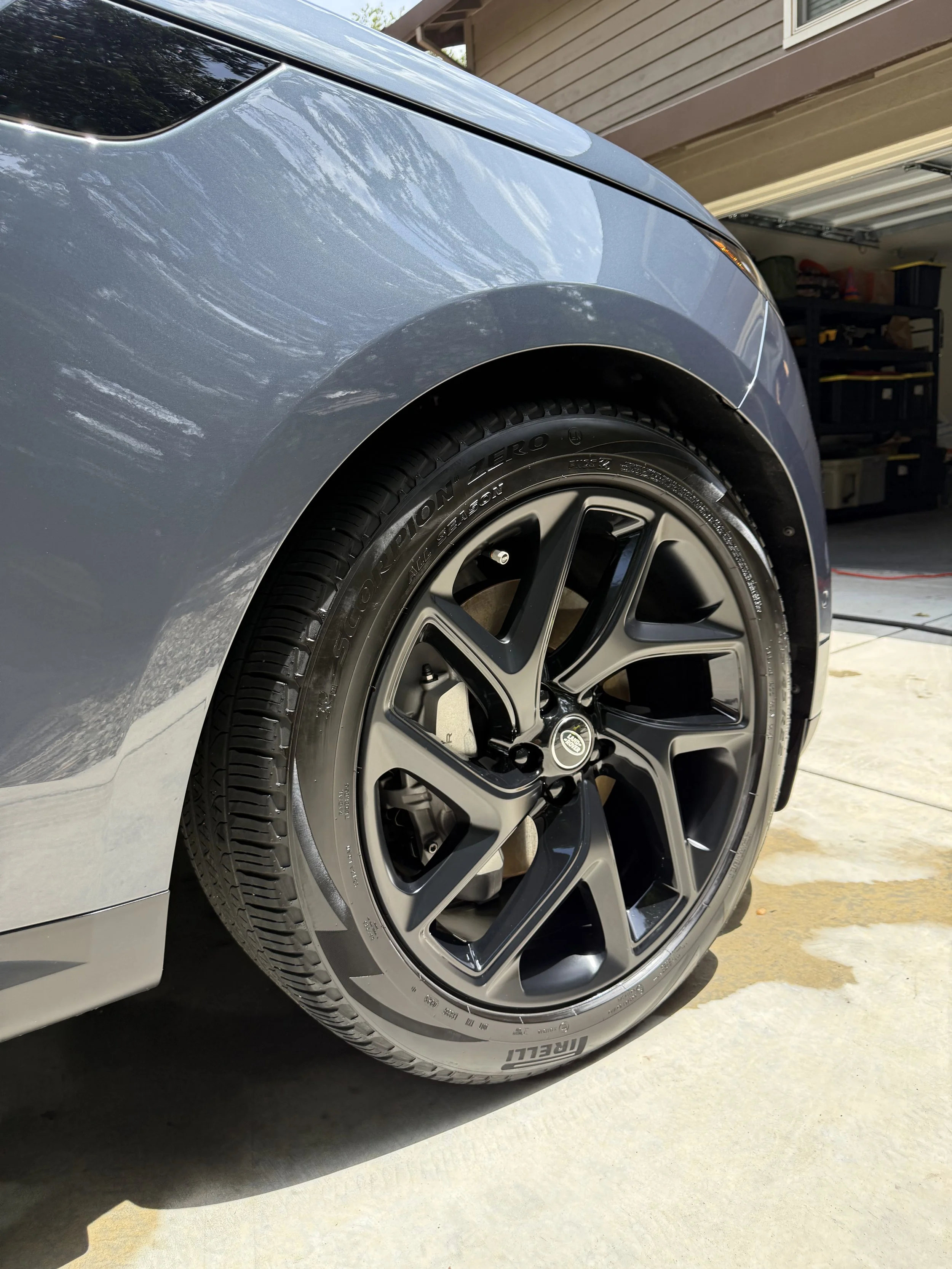 Close-up of a silver car's front wheel and fender, with a garage and storage shelves in the background.