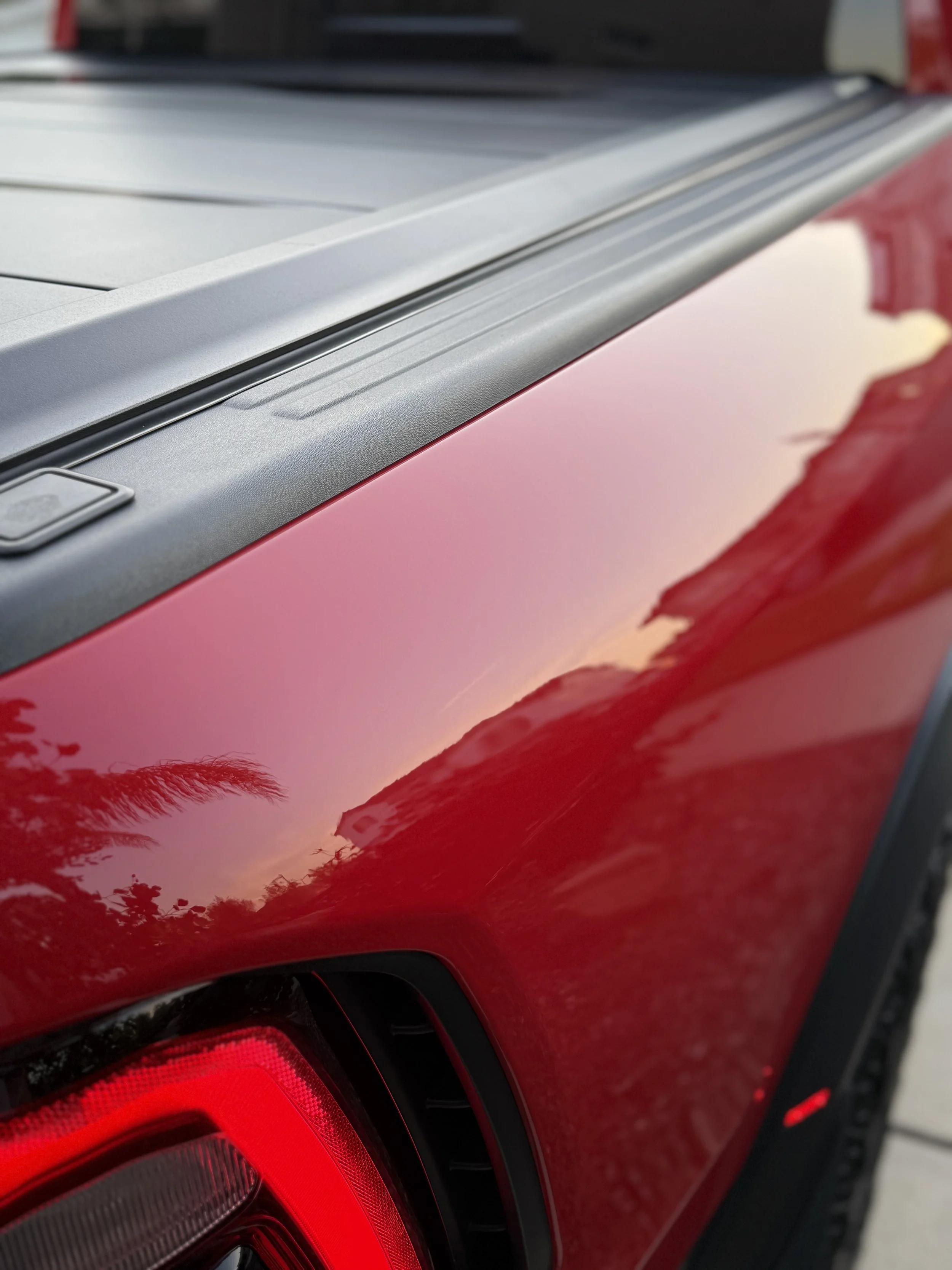 Close-up of a red pickup truck's tailgate and rear taillight, with reflections of the sky and trees on the shiny surface.