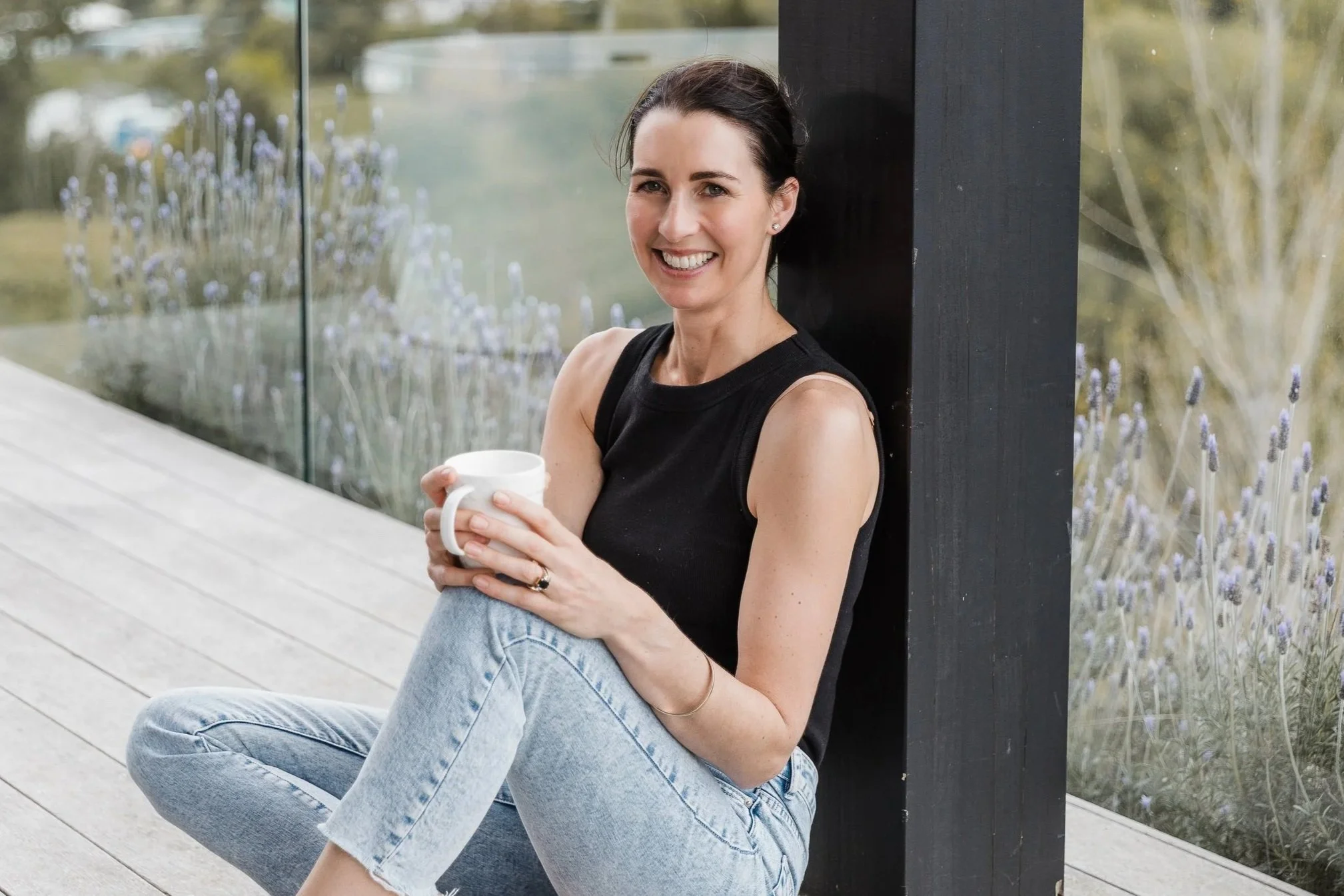 A woman sitting outside on a wooden deck, leaning against a black post, holding a white mug, smiling at the camera, with a garden of lavender flowers in the background.