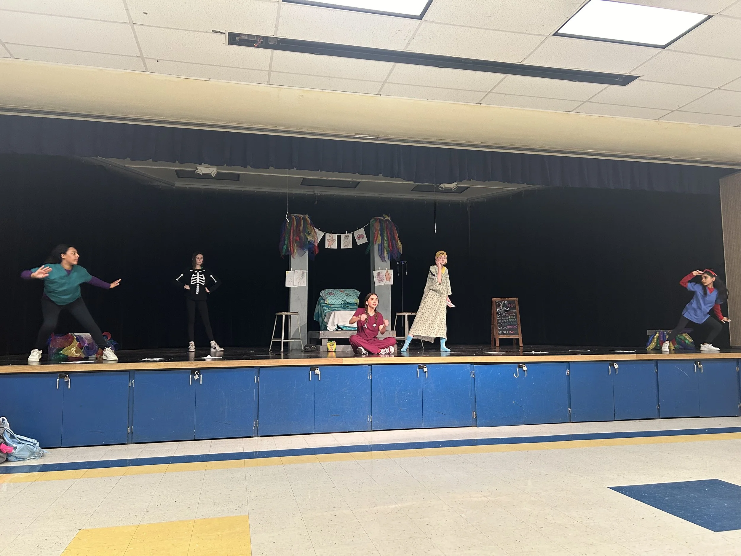 Children performing a play or skit on a stage in a school auditorium with costumes and props.
