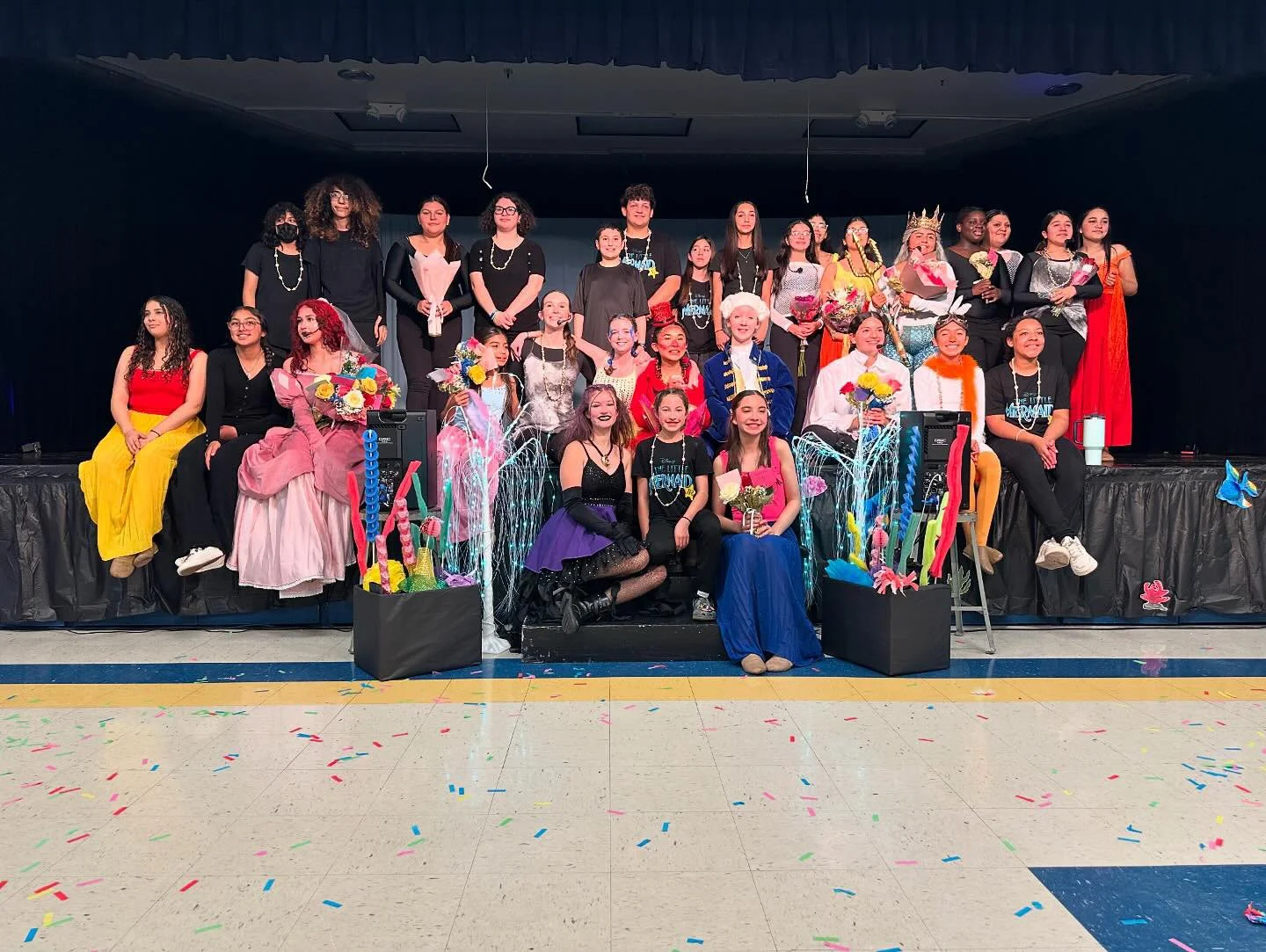 Group photo of children and adults on stage after a performance, some dressed in colorful costumes, with flowers and decorations in front, confetti on the floor, and a black curtain backdrop.