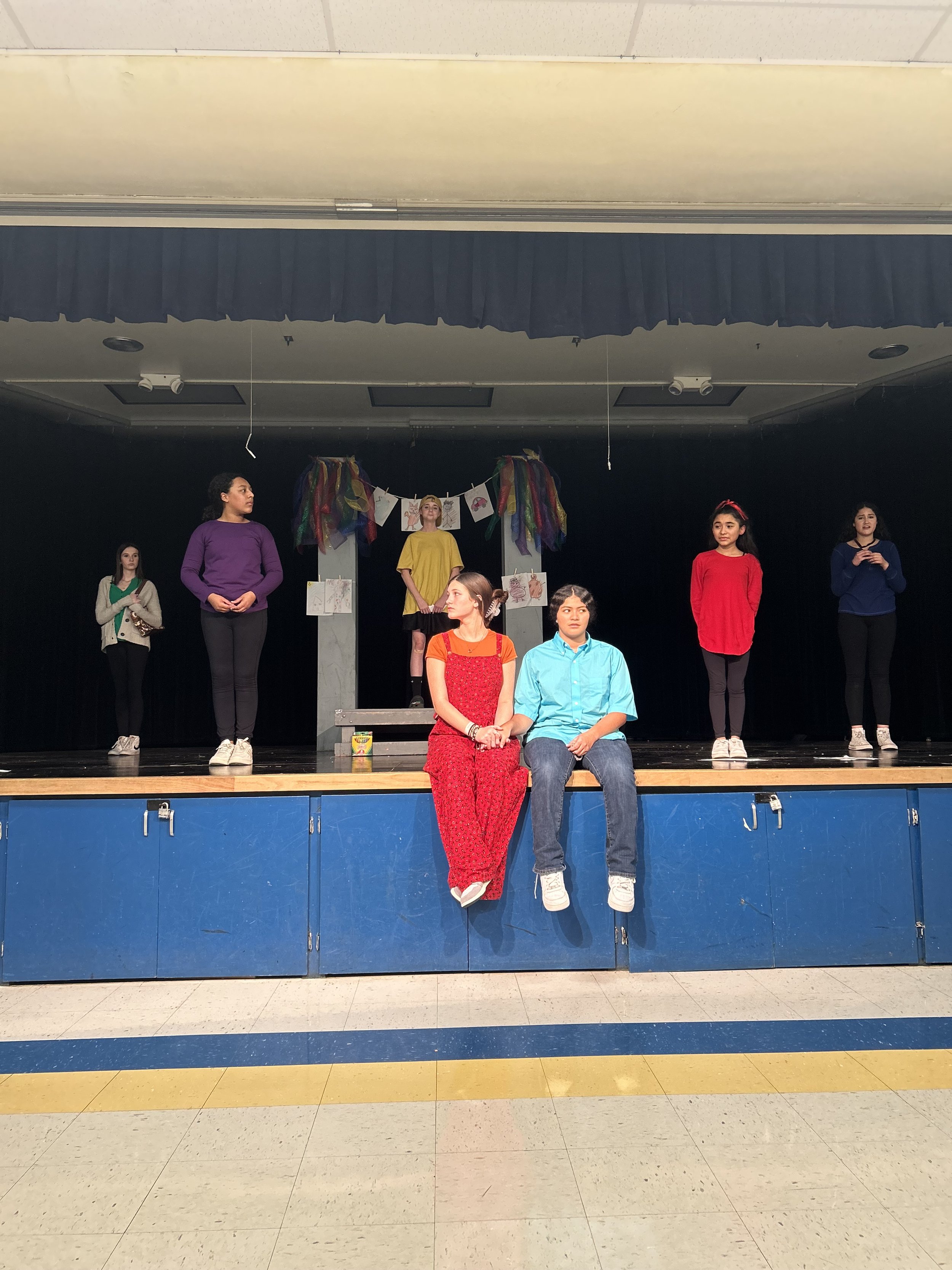 Young girls performing a play on stage, with two sitting on the edge of the stage holding hands, while others stand behind them, and a colorful decorated backdrop.