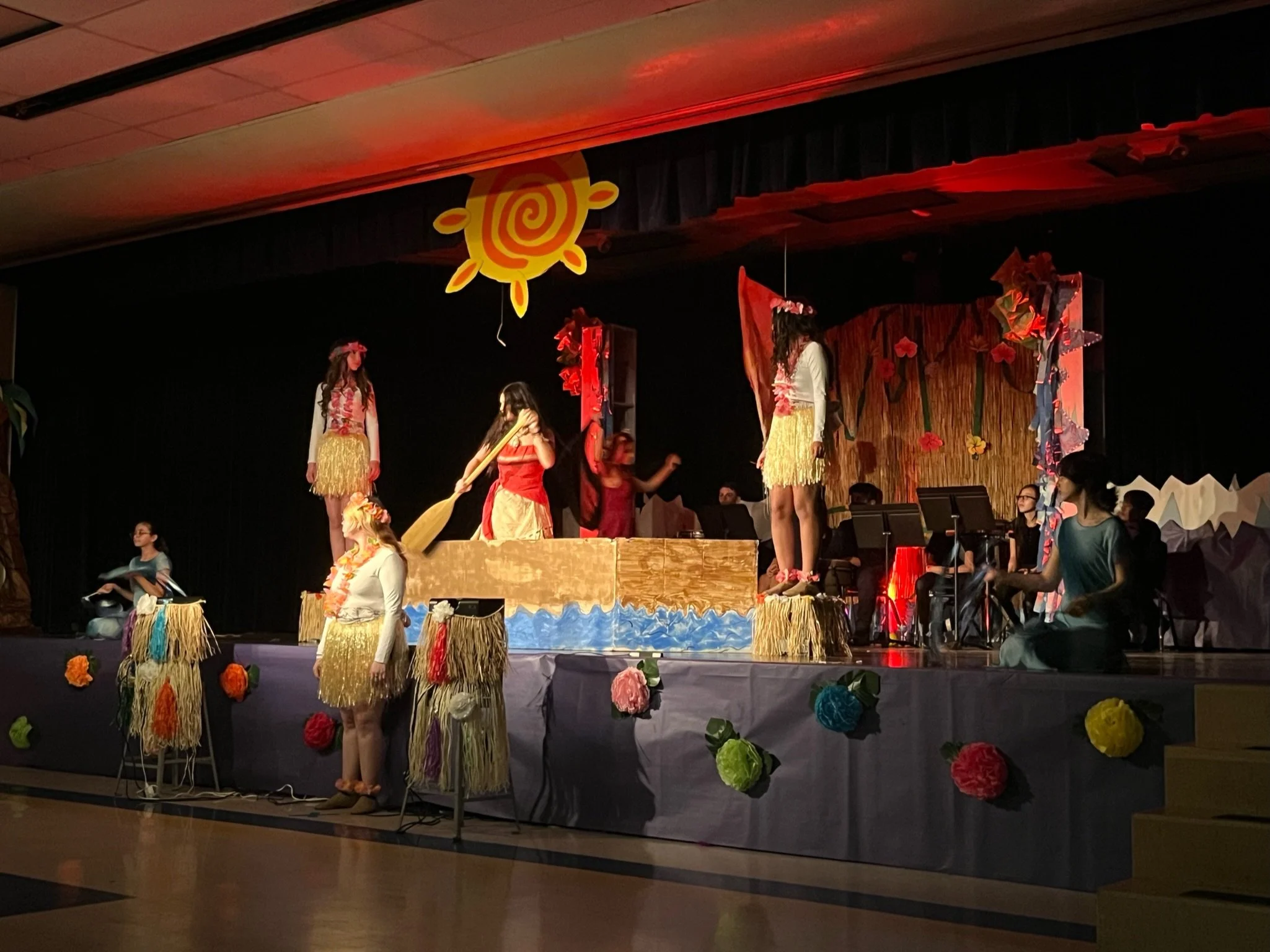 Students performing a play on stage with a Hawaiian theme, dressed in costumes with grass skirts, leis, and floral accessories, surrounded by colorful decorations and a band in the background.