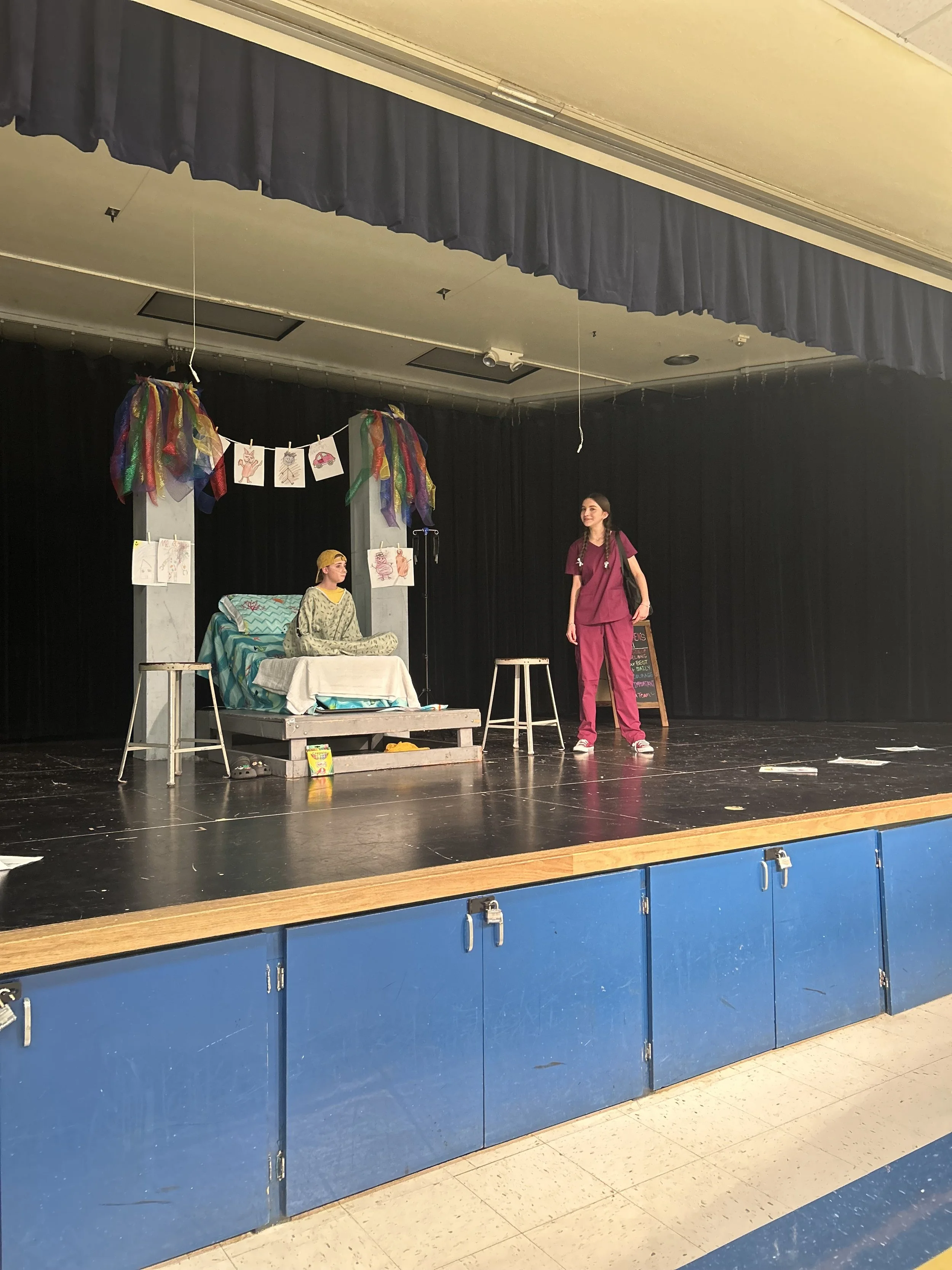 Stage scene from a school play with two young girls in costume. One girl is sitting on a bed, the other standing nearby, with decorations and drawings hanging above the stage.
