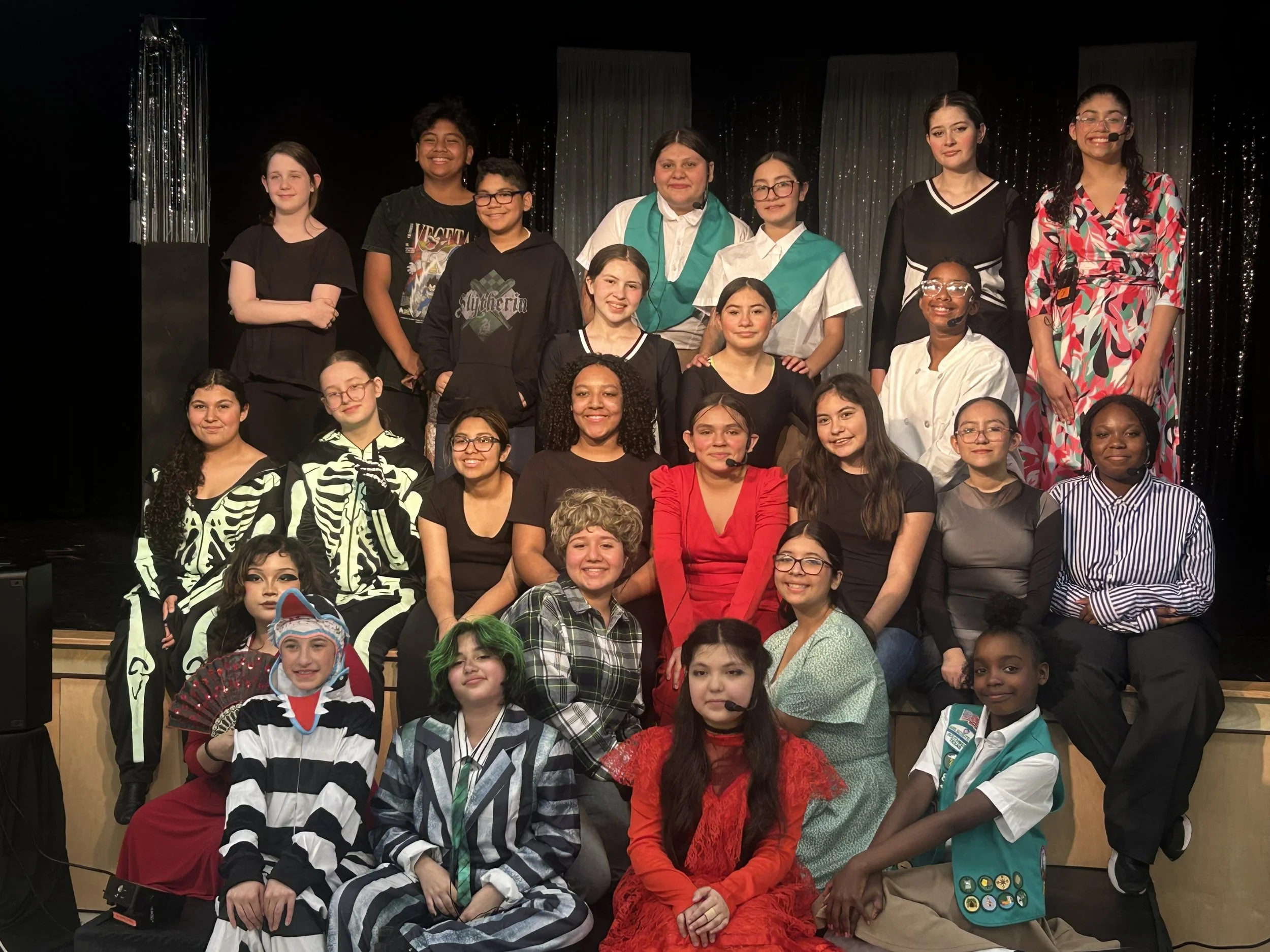 Group of young girls and women on stage for a theatrical performance or event, some in costumes and others in casual or formal attire, against a backdrop of black curtains with silver shimmering details.