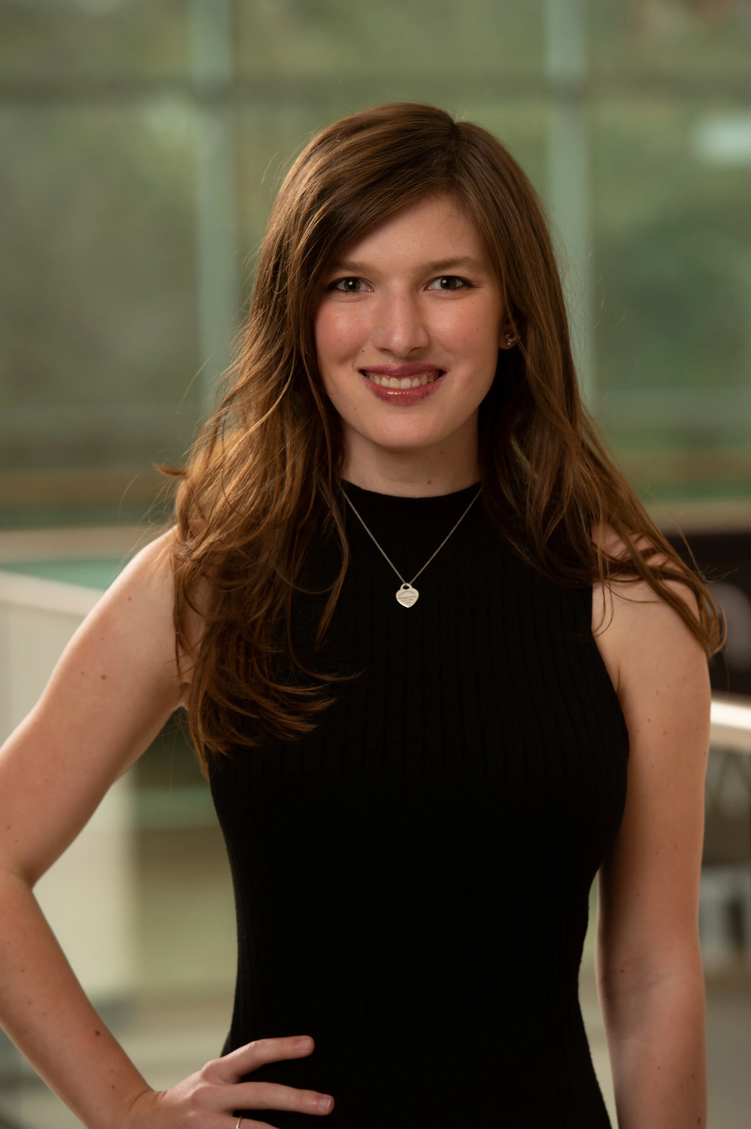 A young woman with long, wavy auburn hair is smiling and posing in a black sleeveless top, standing indoors with a blurred background.