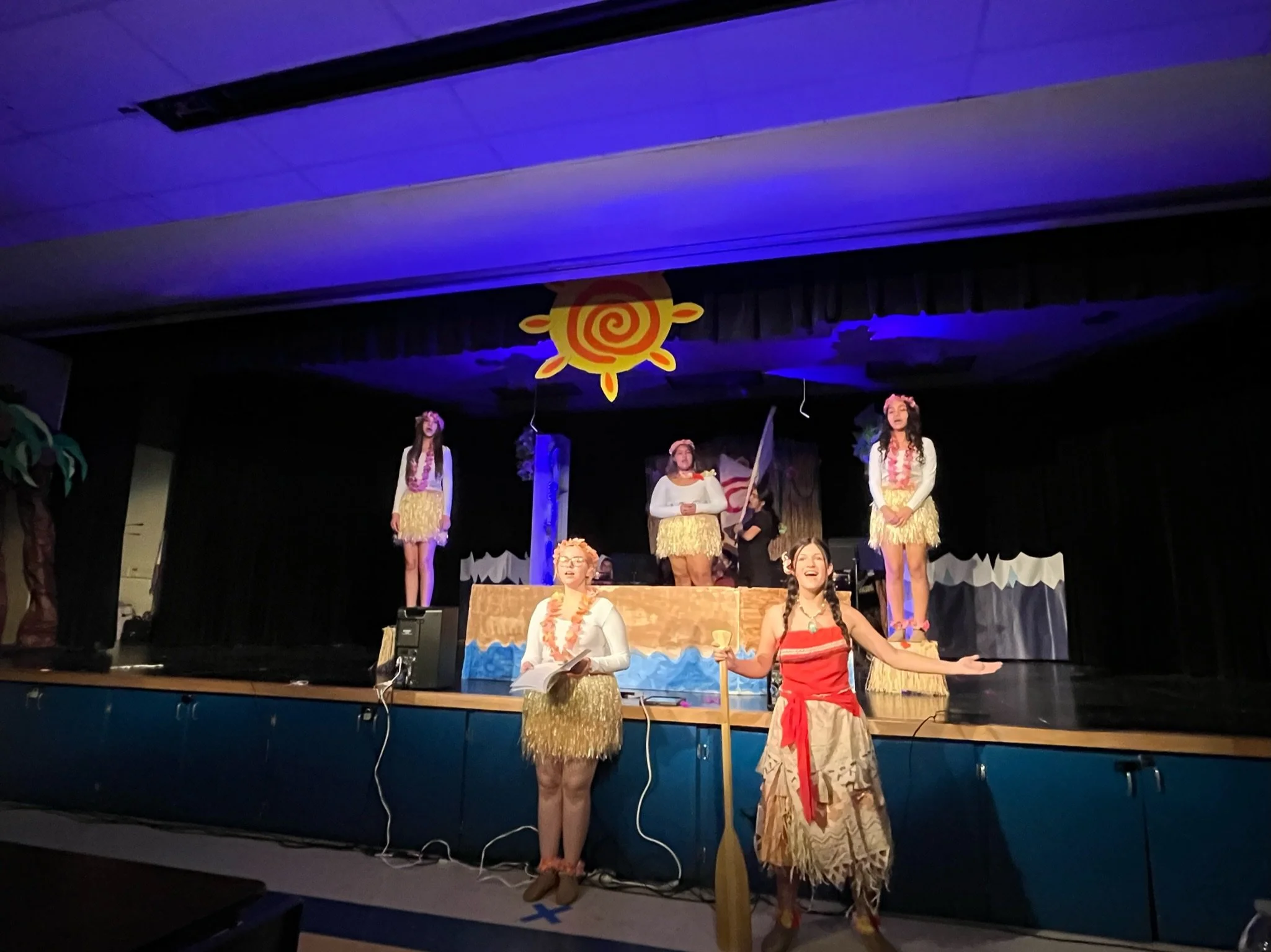 People dressed in Hawaiian-themed costumes on a stage with a beach and sun decorations, performing a play or skit.