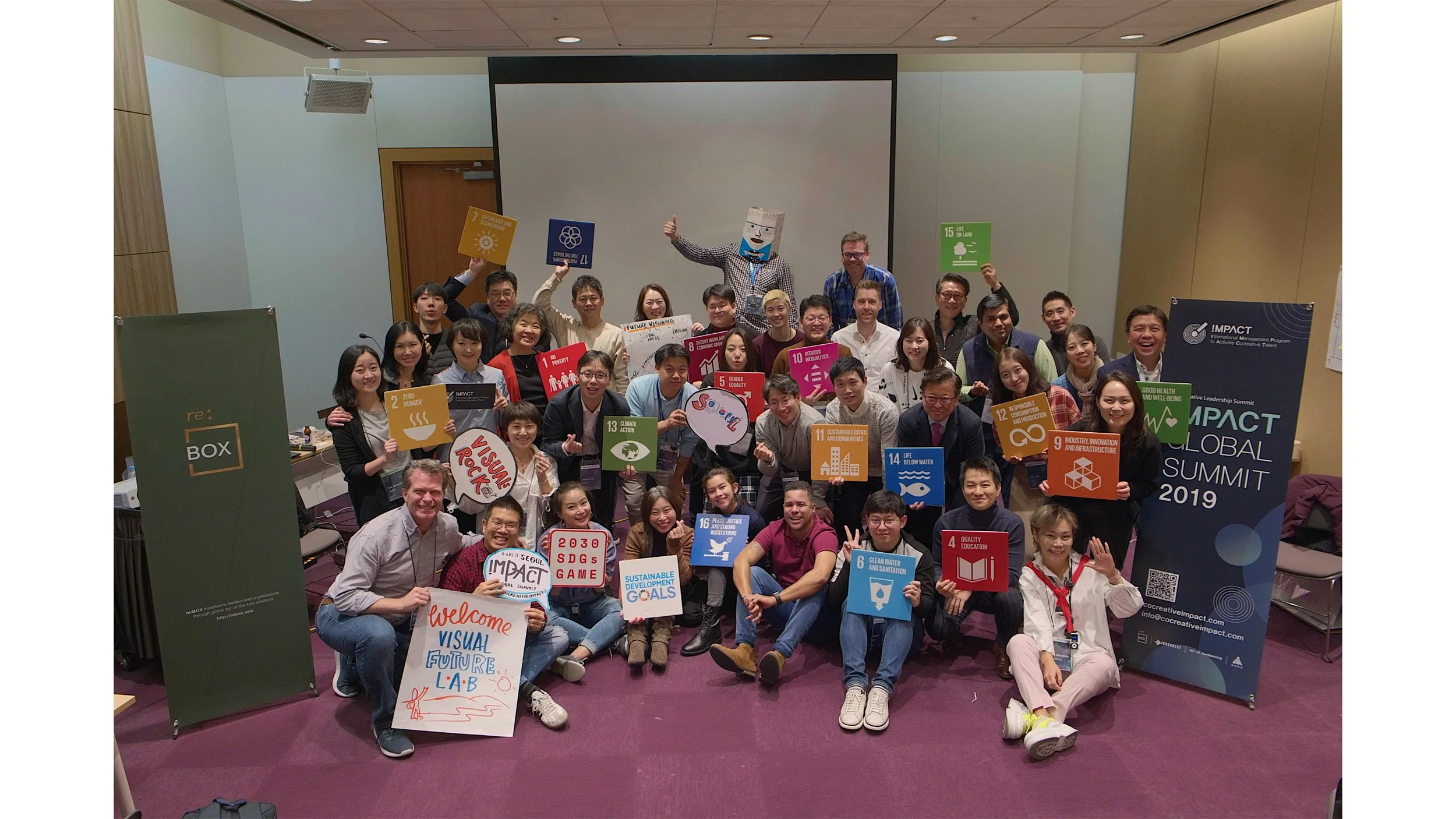 Group of people at Impact Global Summit 2019 holding signs related to sustainable development goals, some smiling and waving, in a conference room with banners and a large screen.