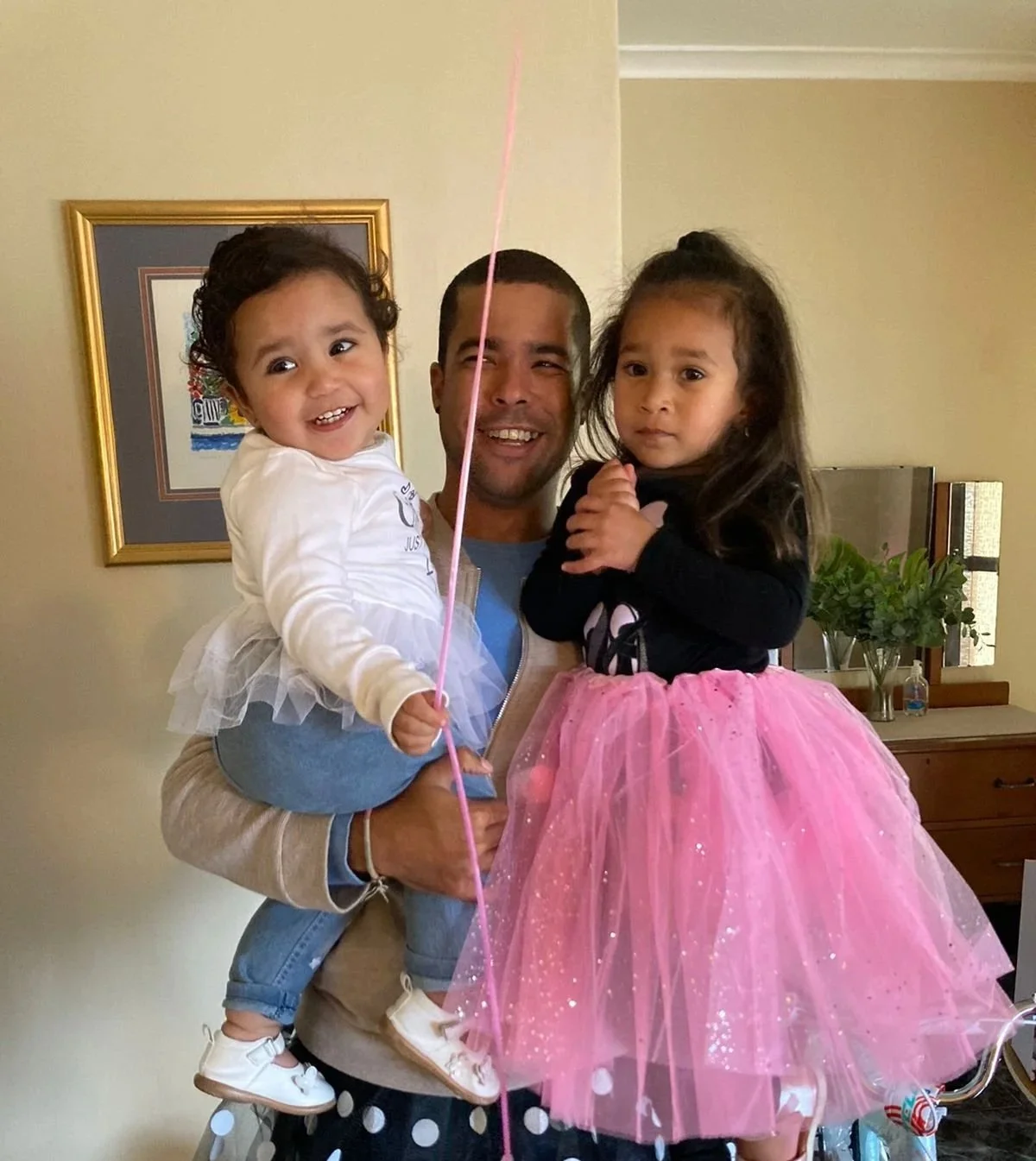 Romano Theunissen is holding his two young nieces, one on each arm. The girl on the left is smiling, holding a pink balloon, and wearing a white shirt with a tutu. The girl on the right looks serious, wearing a black top and a pink tulle skirt.