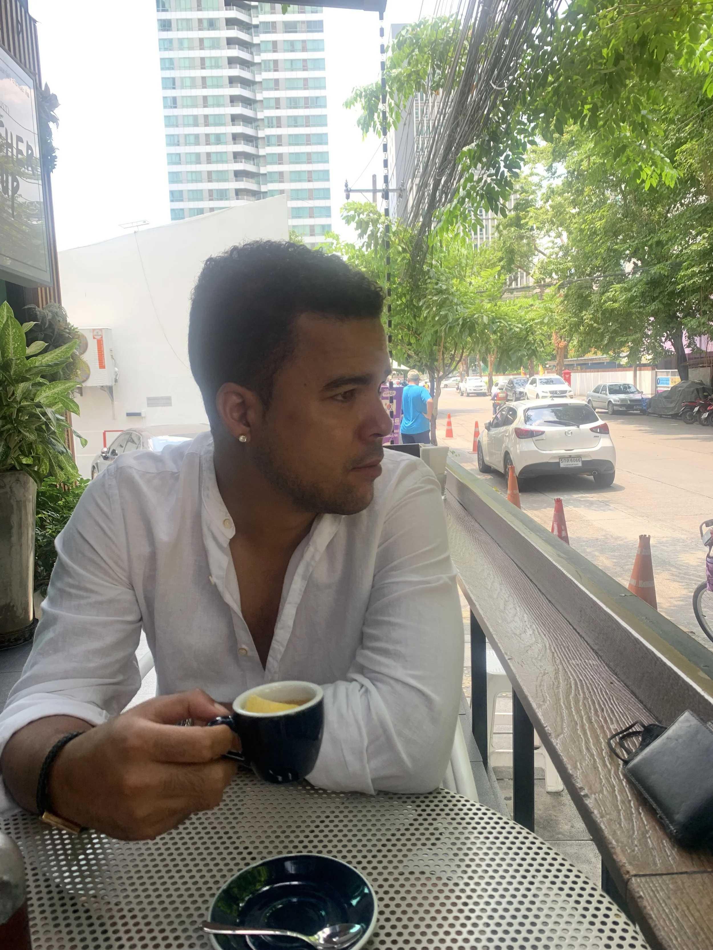 Romano Theunissen in a white shirt sitting at an outdoor cafe table, holding a cup of coffee, with a city street view in the background.