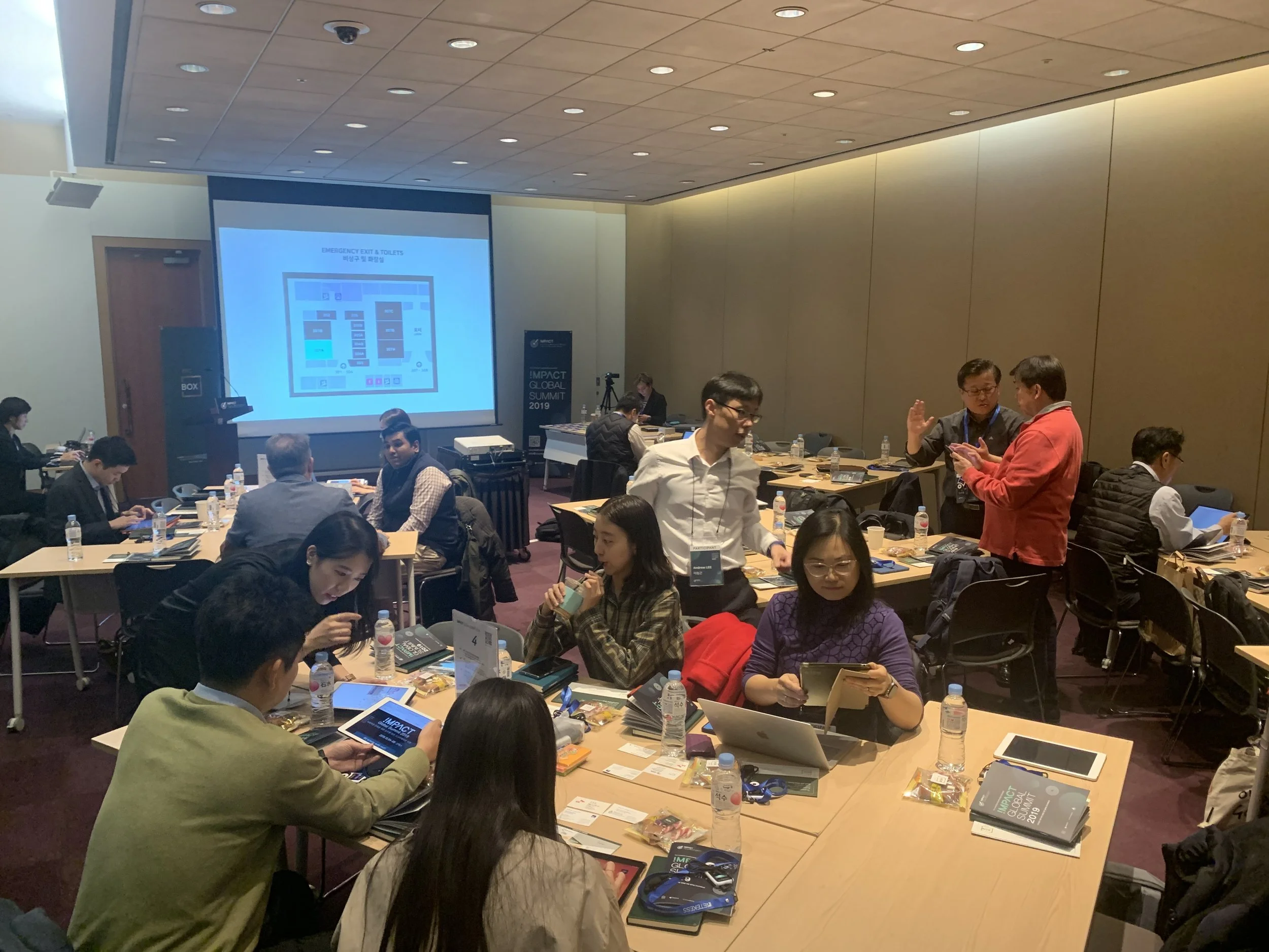 People attending a conference in a room with tables, laptops, bottled water, and conference materials, with a large presentation screen at the front.
