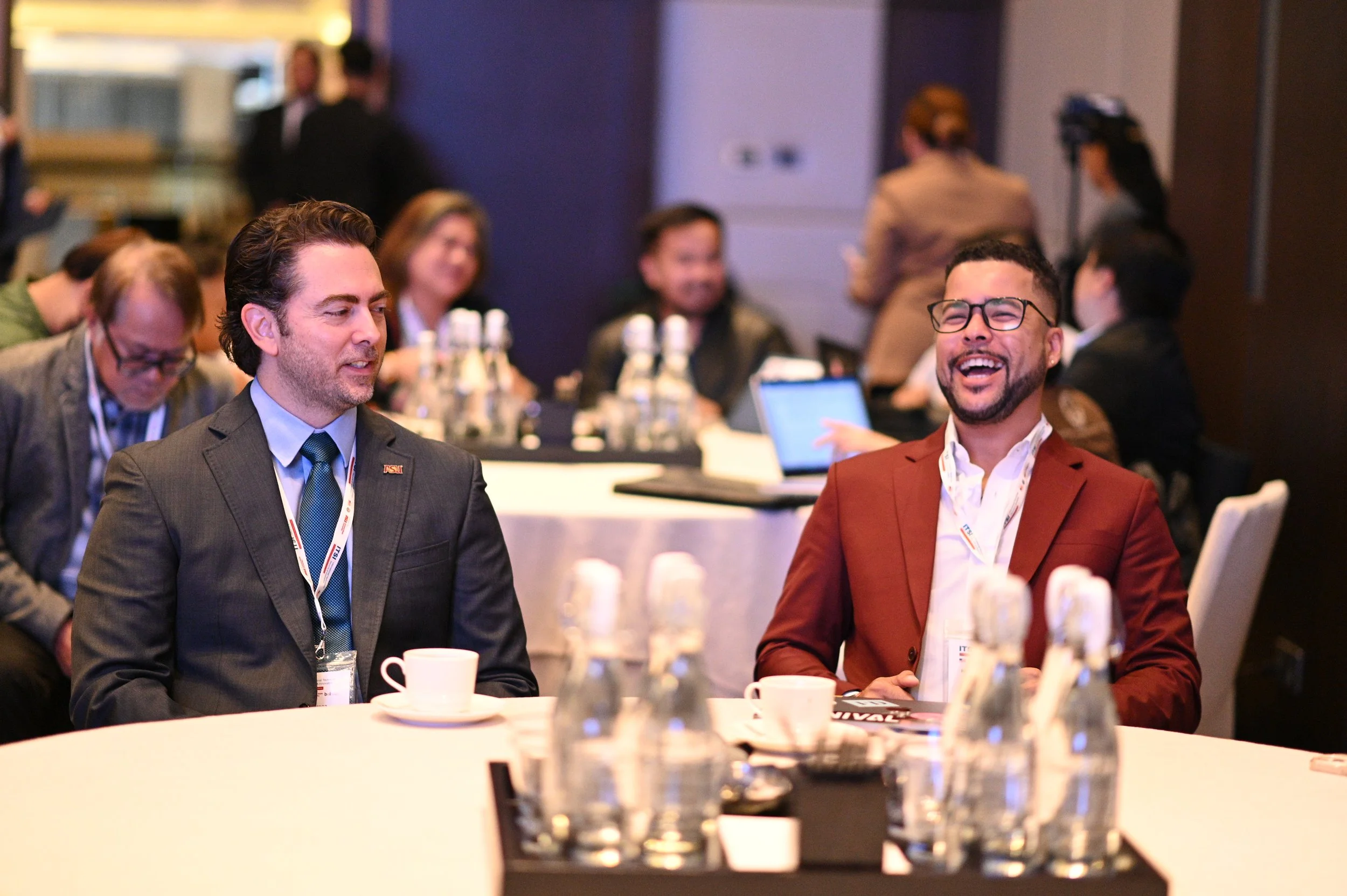 Two men in suits sitting at a round conference table, smiling and laughing, with water bottles, coffee cups, and conference materials in front of them, surrounded by other conference attendees in a bright room.