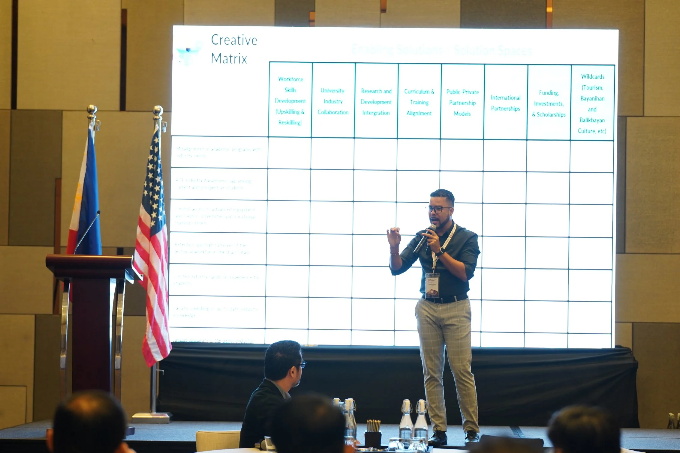 Romano Theunissen speaking into a microphone on stage at a conference, with a large presentation slide titled 'Creative Matrix' and American flags nearby.