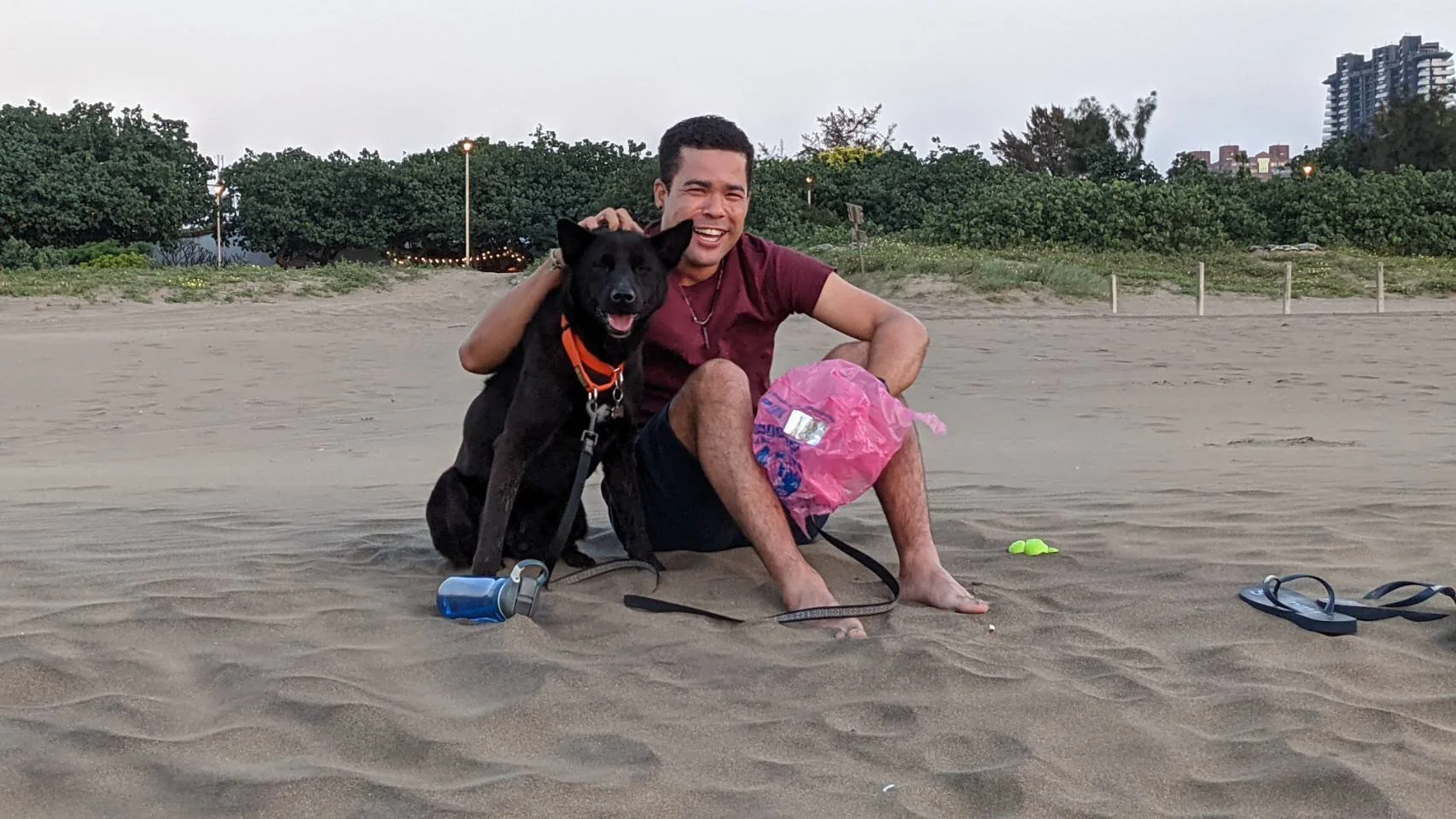 Romano Theunissen and his black dog sitting on a sandy beach smiling at the camera, with trees and buildings in the background.