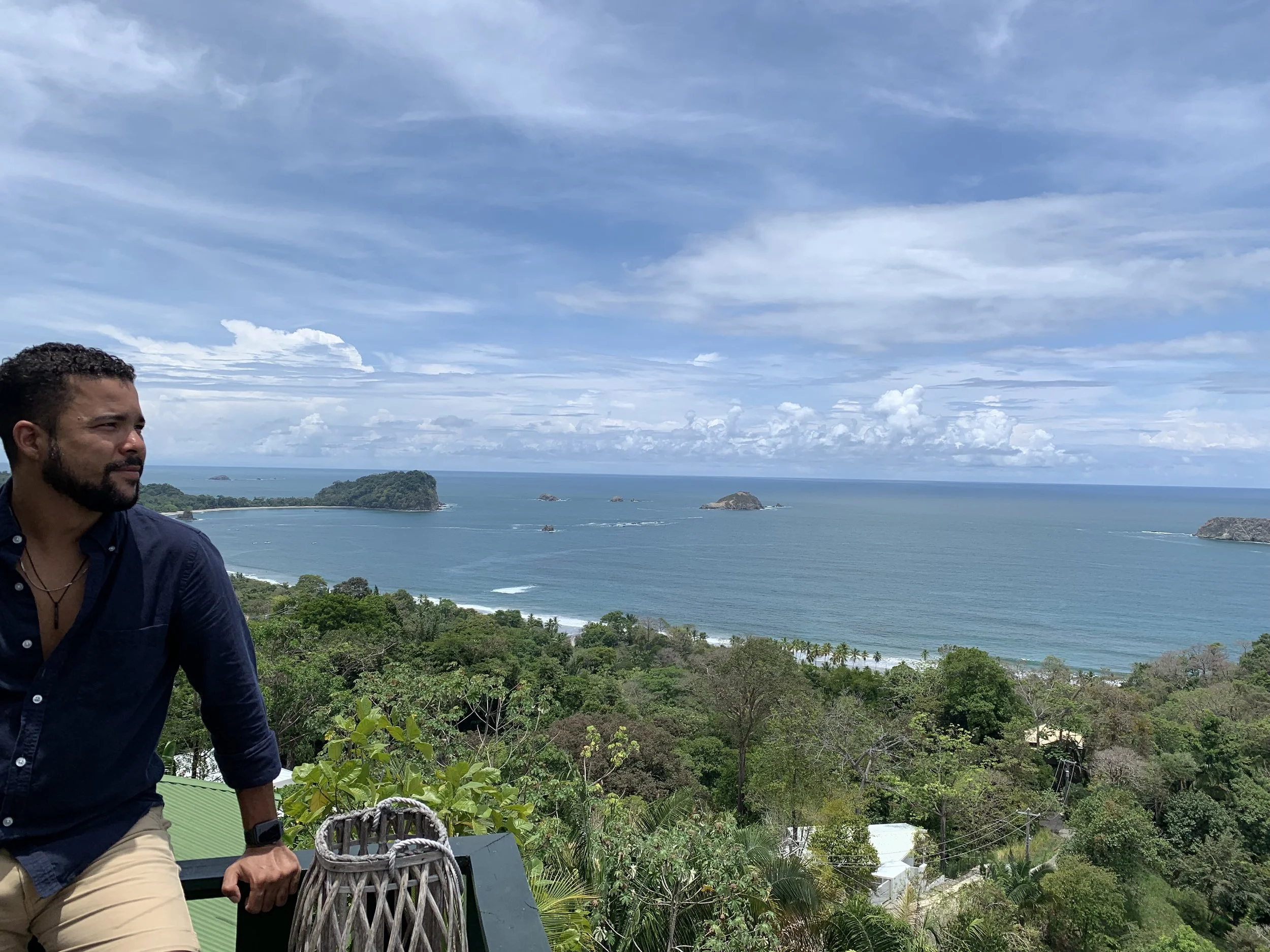 Romano Theunissen with a beard and dark hair, wearing a dark blue shirt and beige shorts, sitting on a railing overlooking a coastal landscape with trees, islands, and the ocean under a partly cloudy sky in Costa Rica.