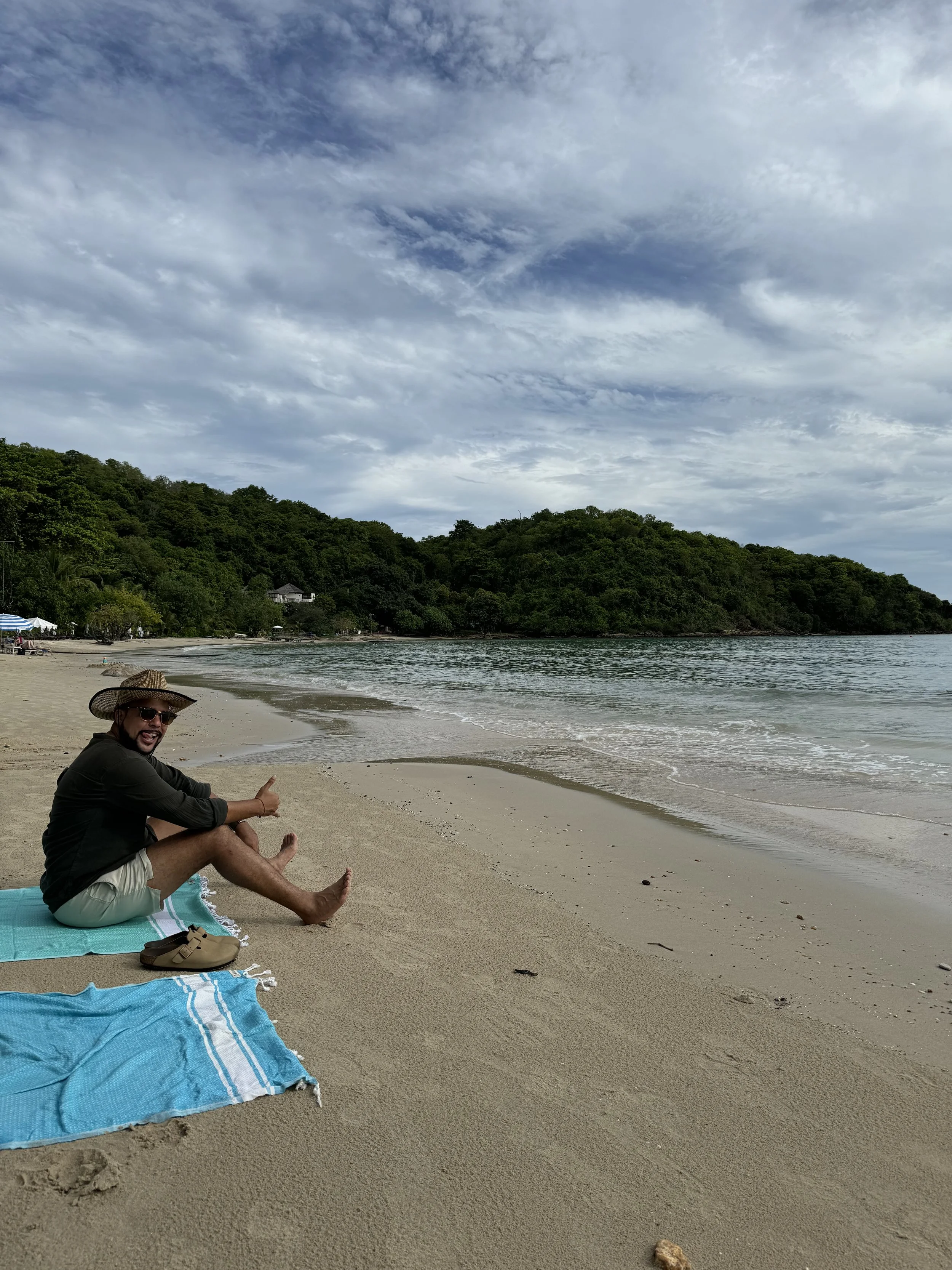 Romano Theunissen sitting on a beach towel with a hat and sunglasses, giving a thumbs-up, with a body of water and green hills in the background under a cloudy sky.