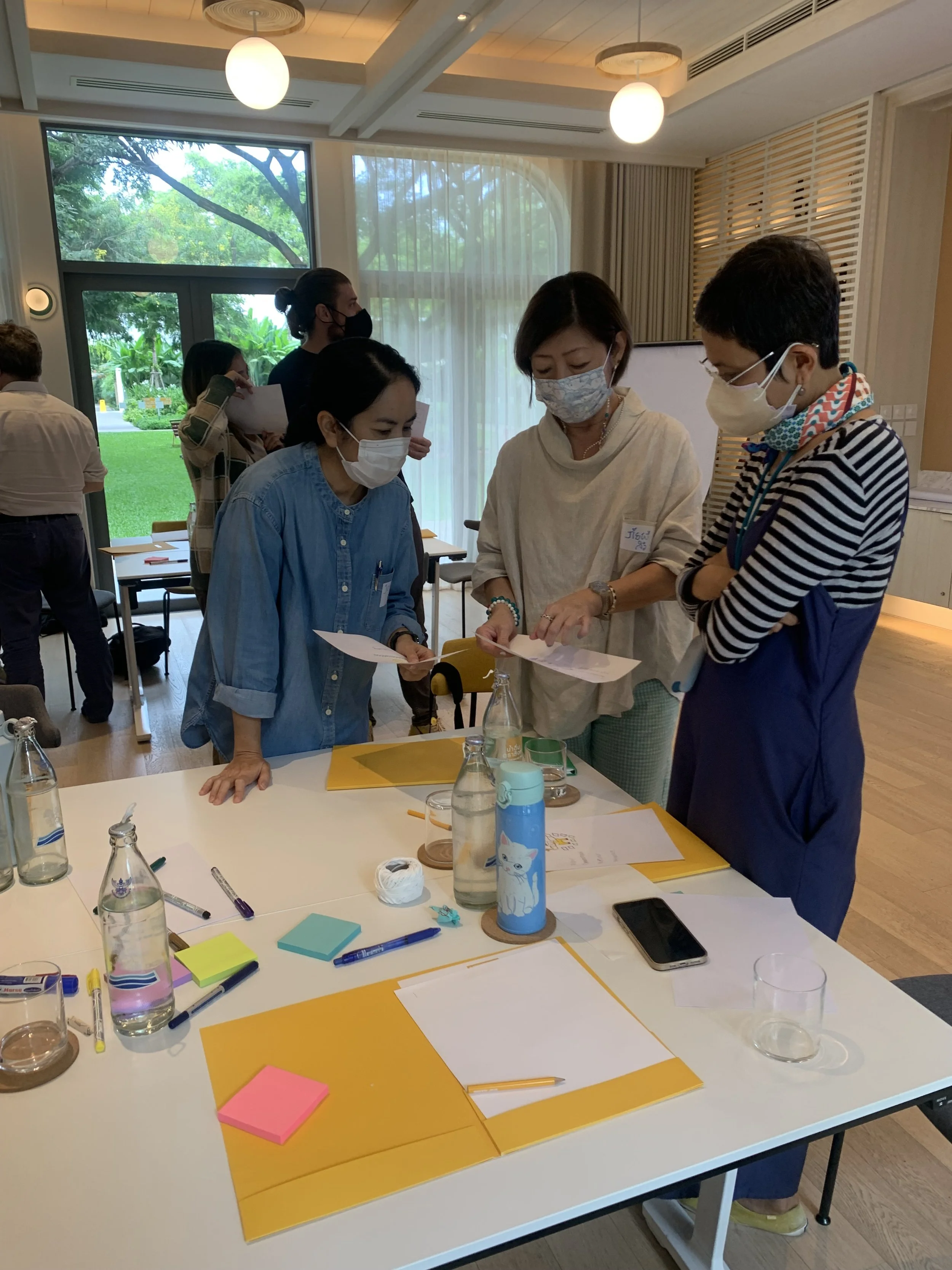 Four women wearing masks are gathered around a table, discussing and looking at papers during a meeting in a well-lit room with large windows and green outdoor scenery in the background. The table has notebooks, pens, sticky notes, water bottles, and a smartphone.