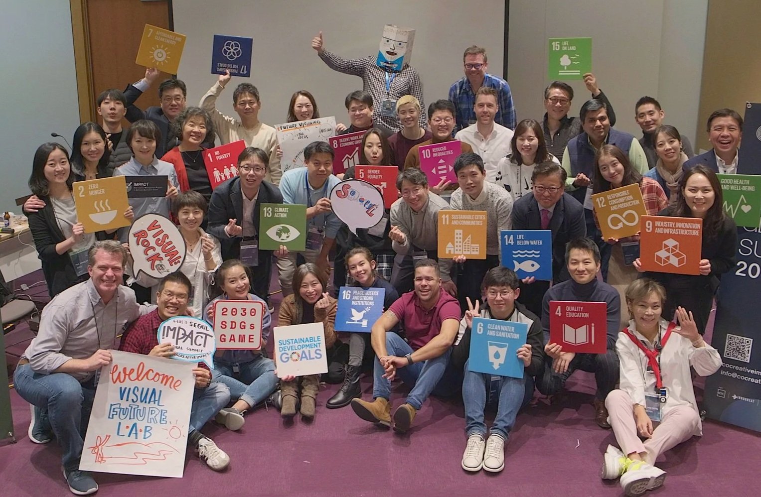 Group of diverse people at a conference holding signs related to the United Nations Sustainable Development Goals, with a person in a Innogreat Box in the background.
