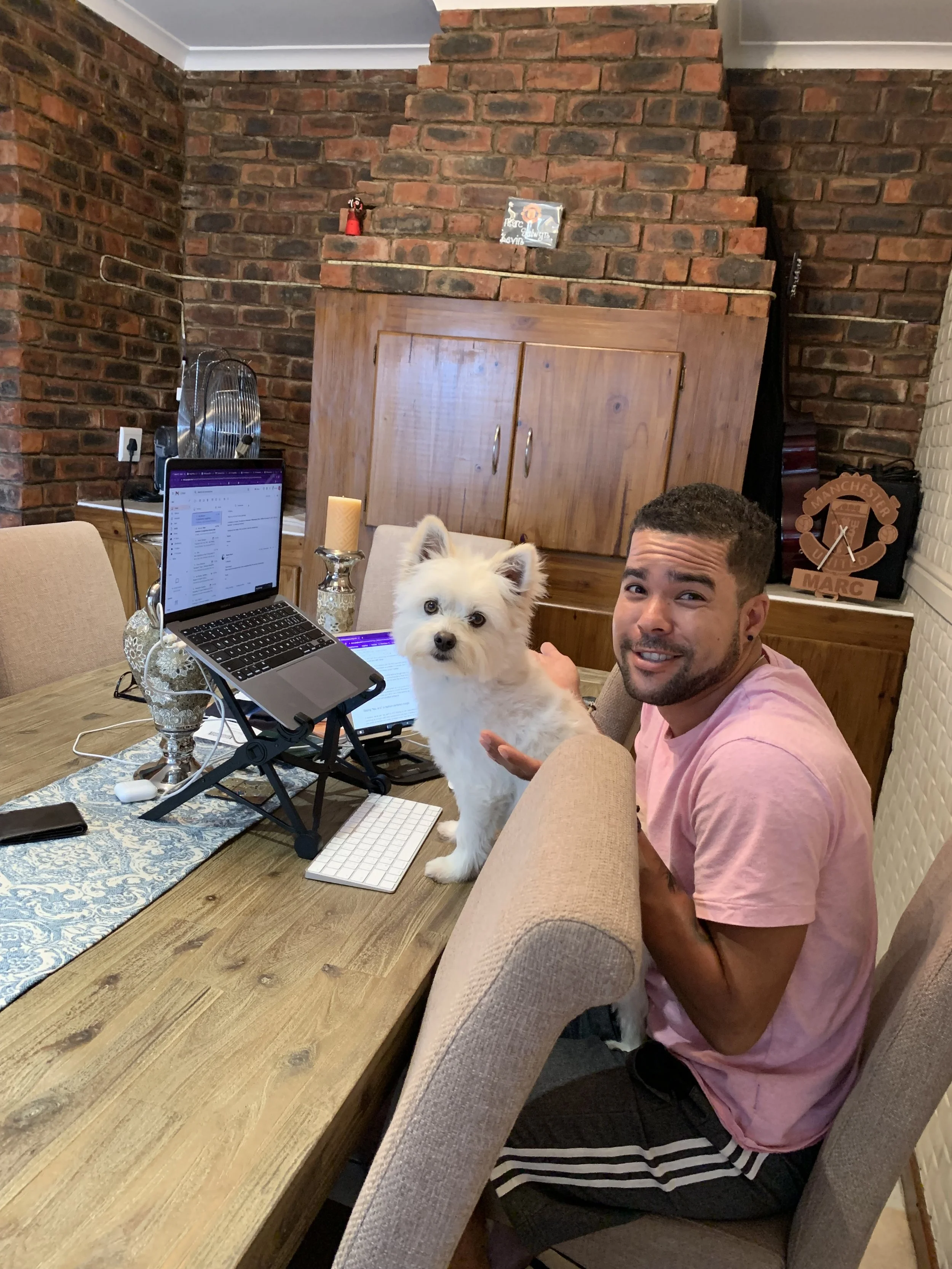 Romano Theunissen is sitting at a dining table with a white dog blocking him from doing work, in a room with brick walls and wooden furniture.