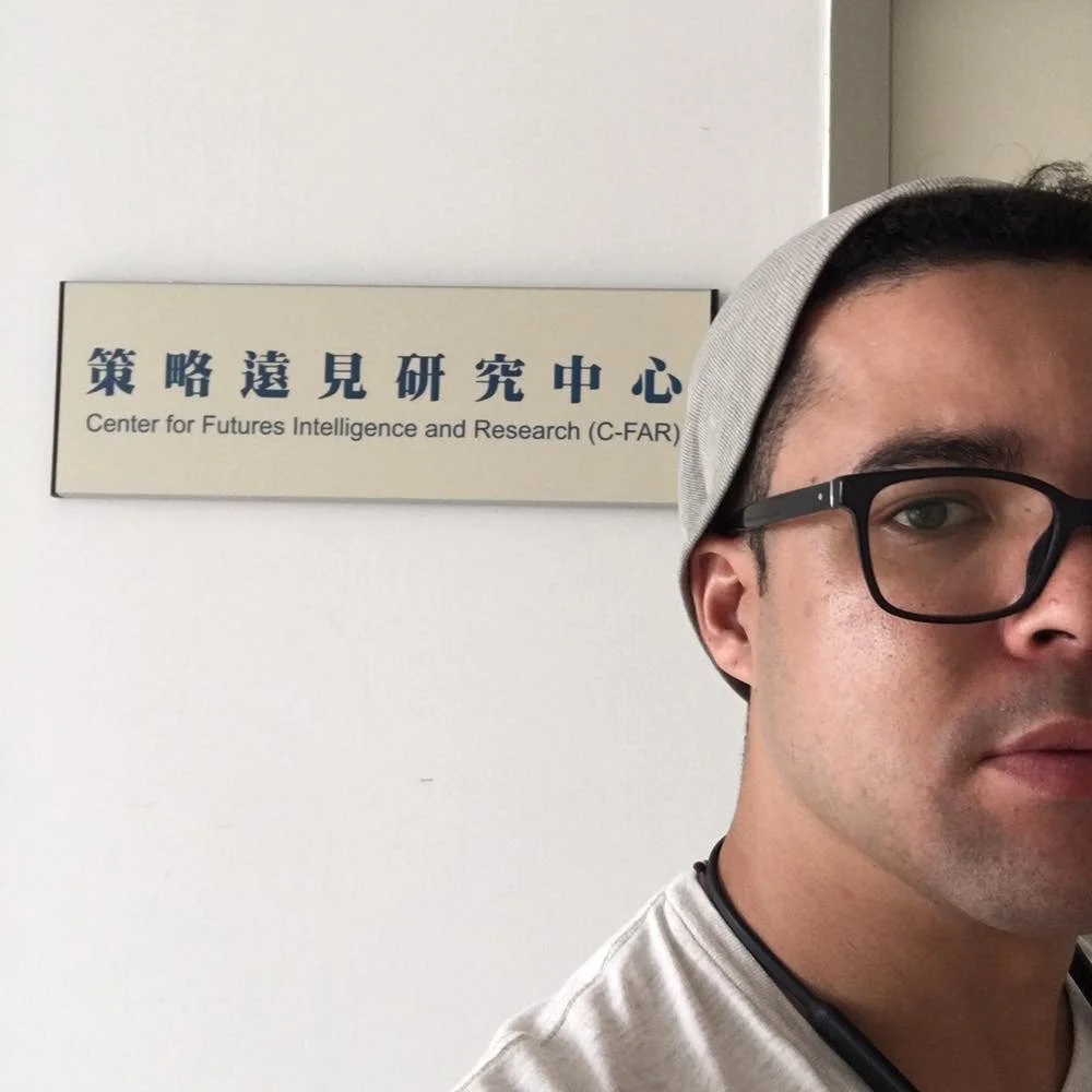 Part of a man wearing glasses and a gray hat, standing in front of a sign that reads 'Center for Futures Intelligence and Research (C-FAR)' in both Chinese and English.
