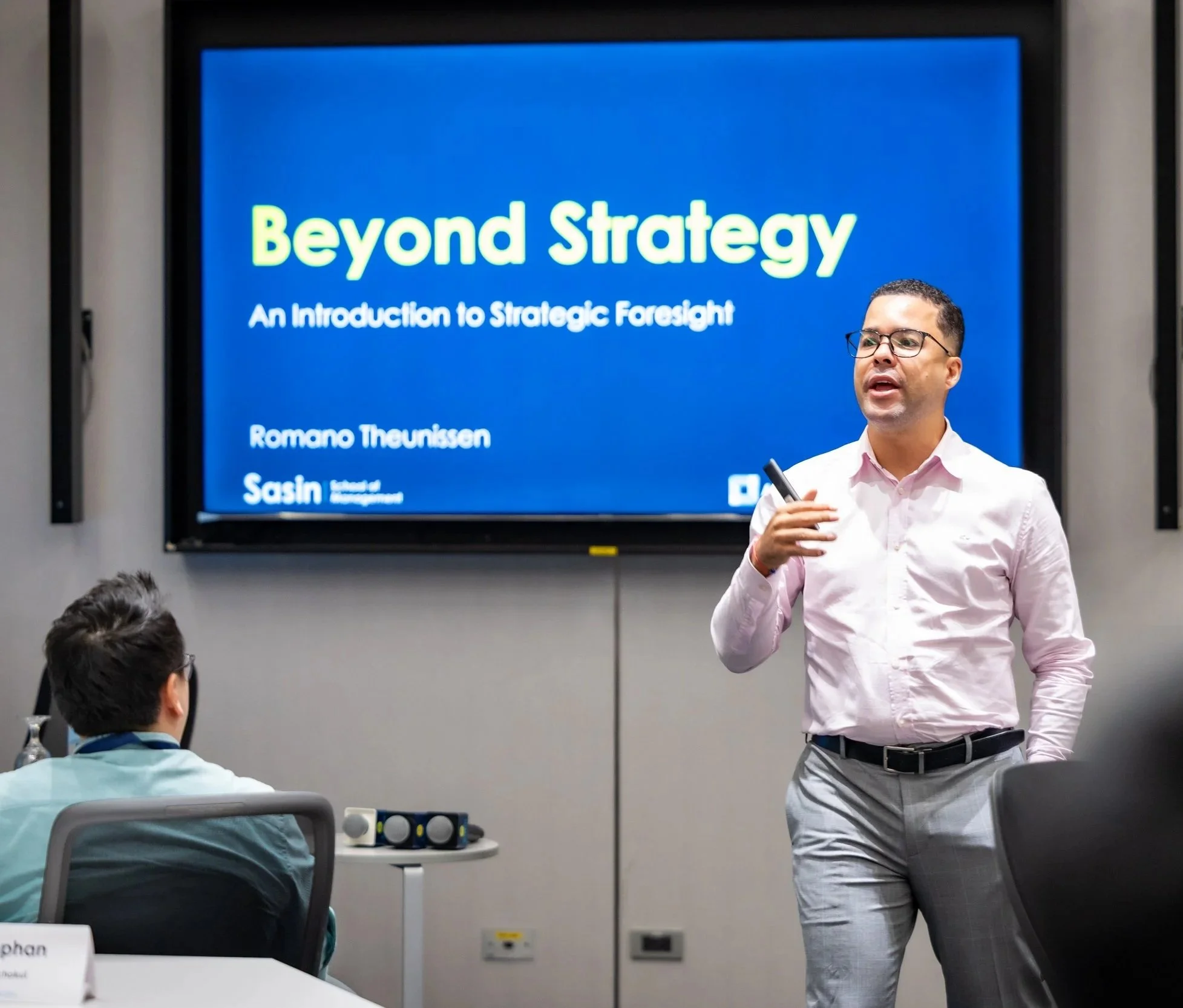 Romano Theunissen teaching a class of MBA students with a large screen behind him displaying a slide titled 'Beyond Strategy'.