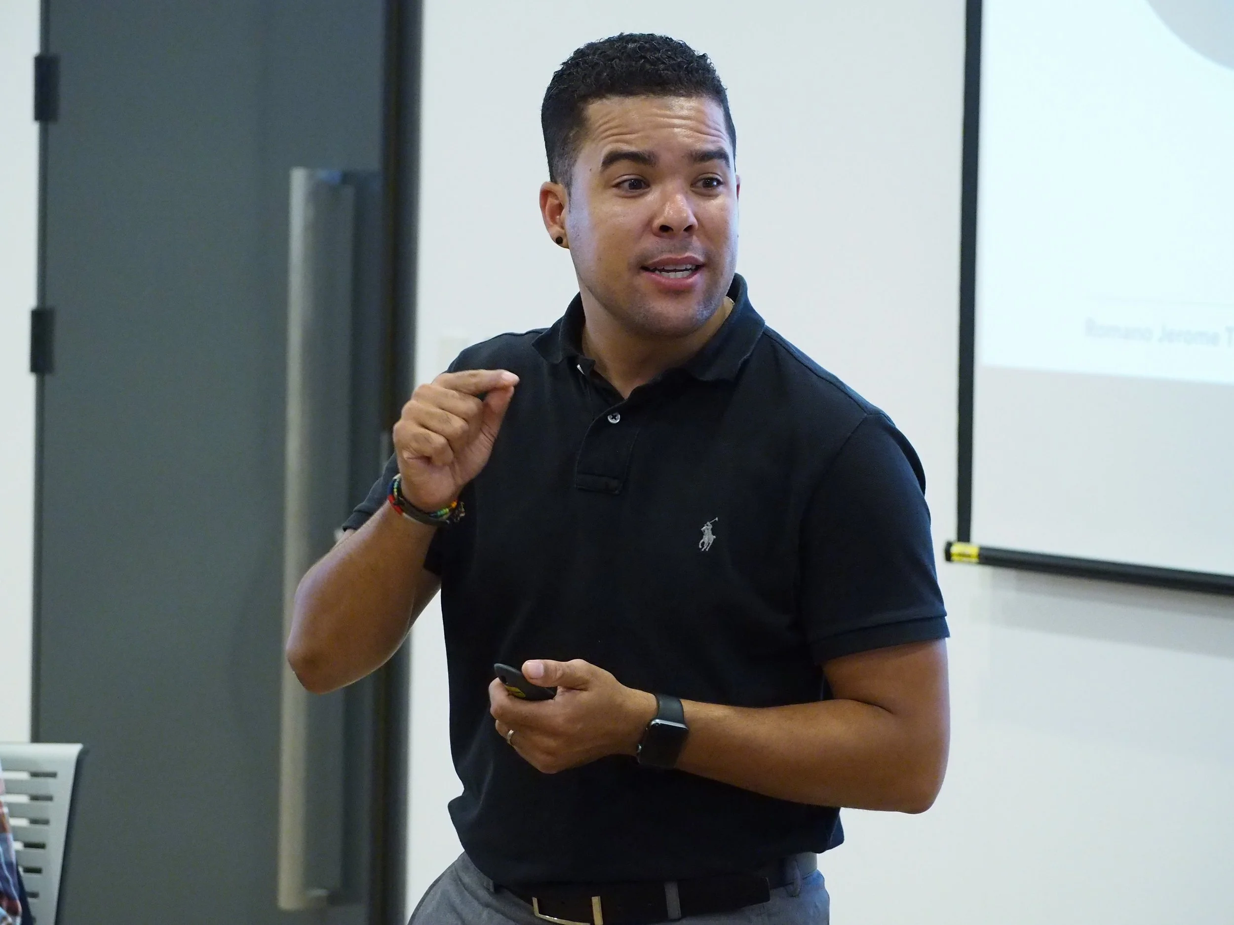 Romano Theunissen in a black polo shirt giving a presentation in a classroom or conference room.