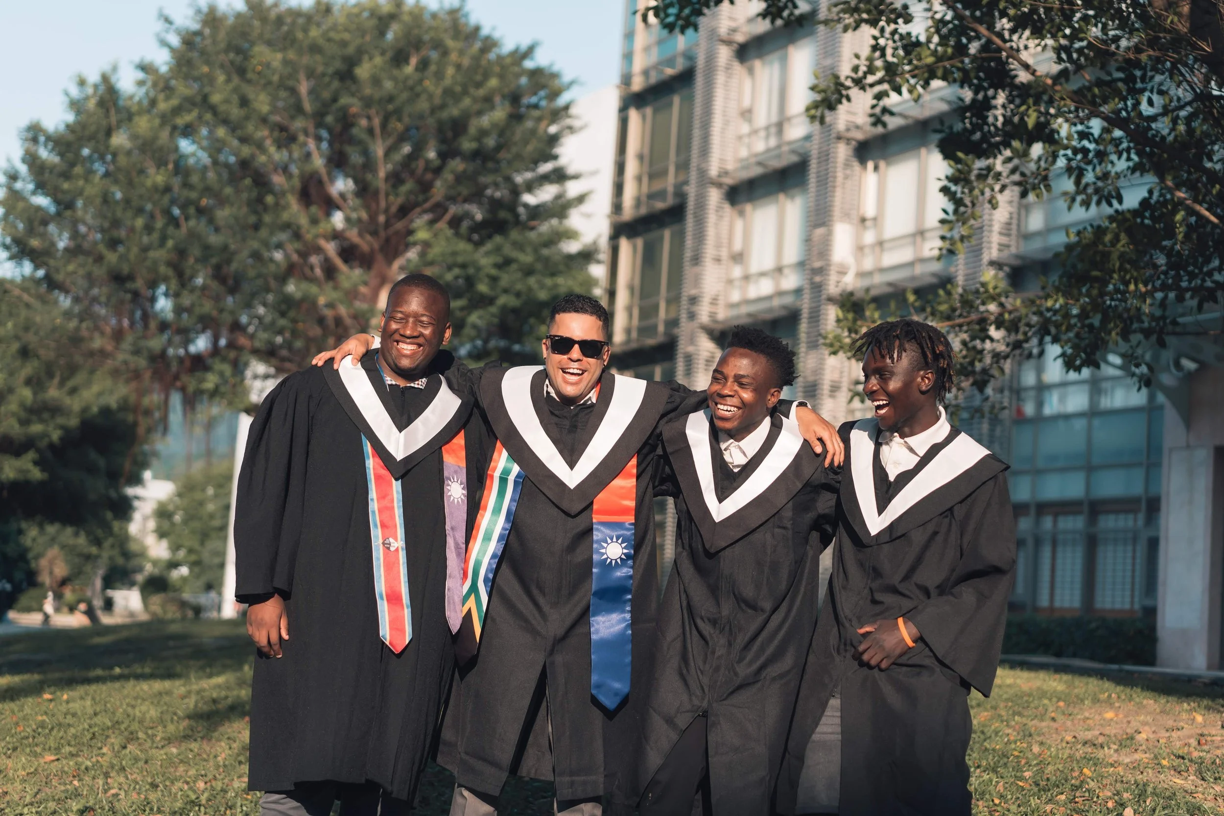 Four diverse young men in graduation robes and academic regalia standing outdoors with arms around each other, smiling and celebrating.